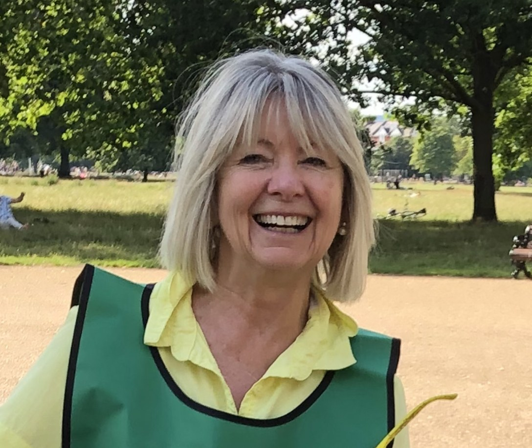 A smiling woman with blonde hair, wearing a yellow shirt and green vest, standing outdoors in a park with trees and grass in the background.