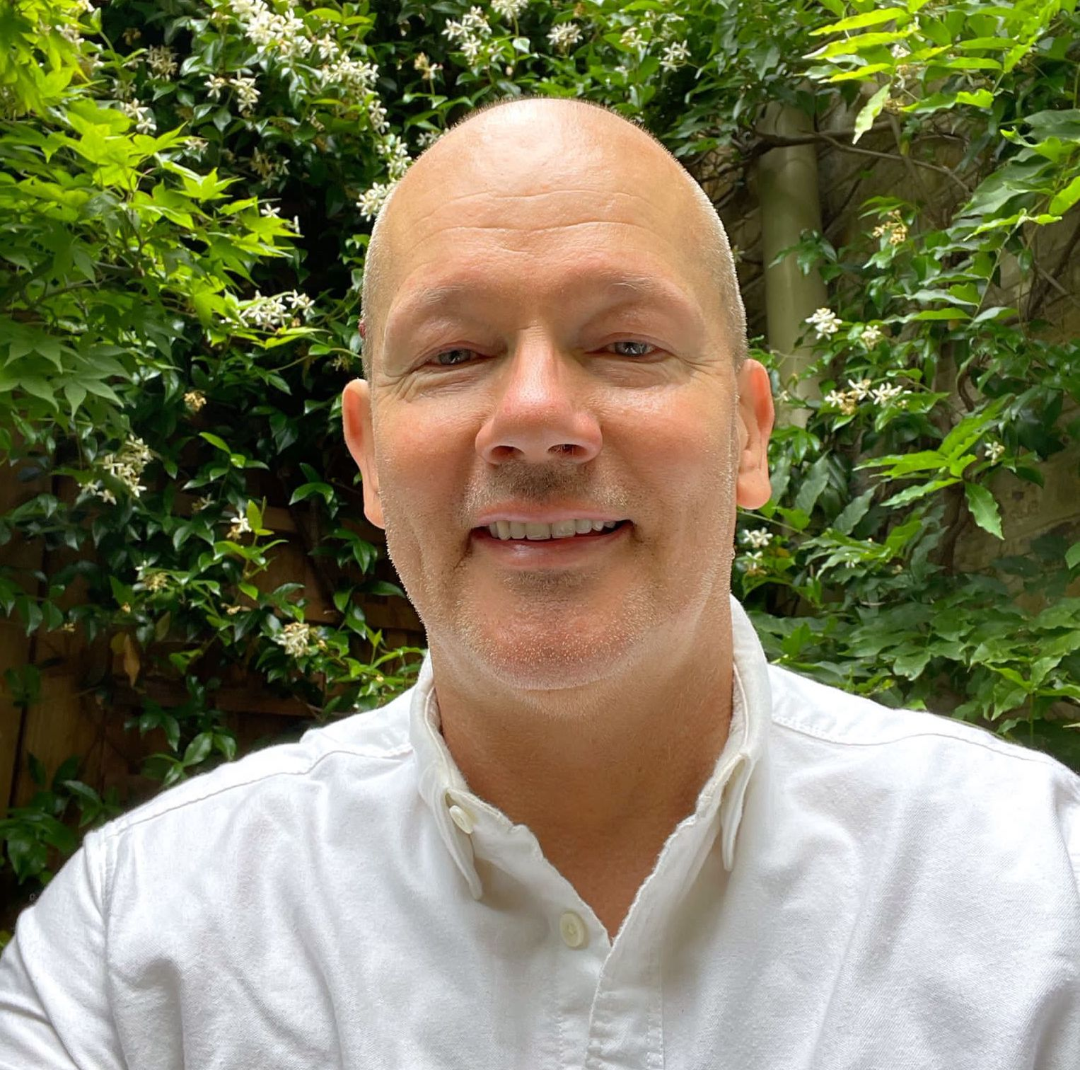 A smiling man with a bald head and light beard, wearing a white shirt, standing outdoors in front of green leafy plants and white flowers.