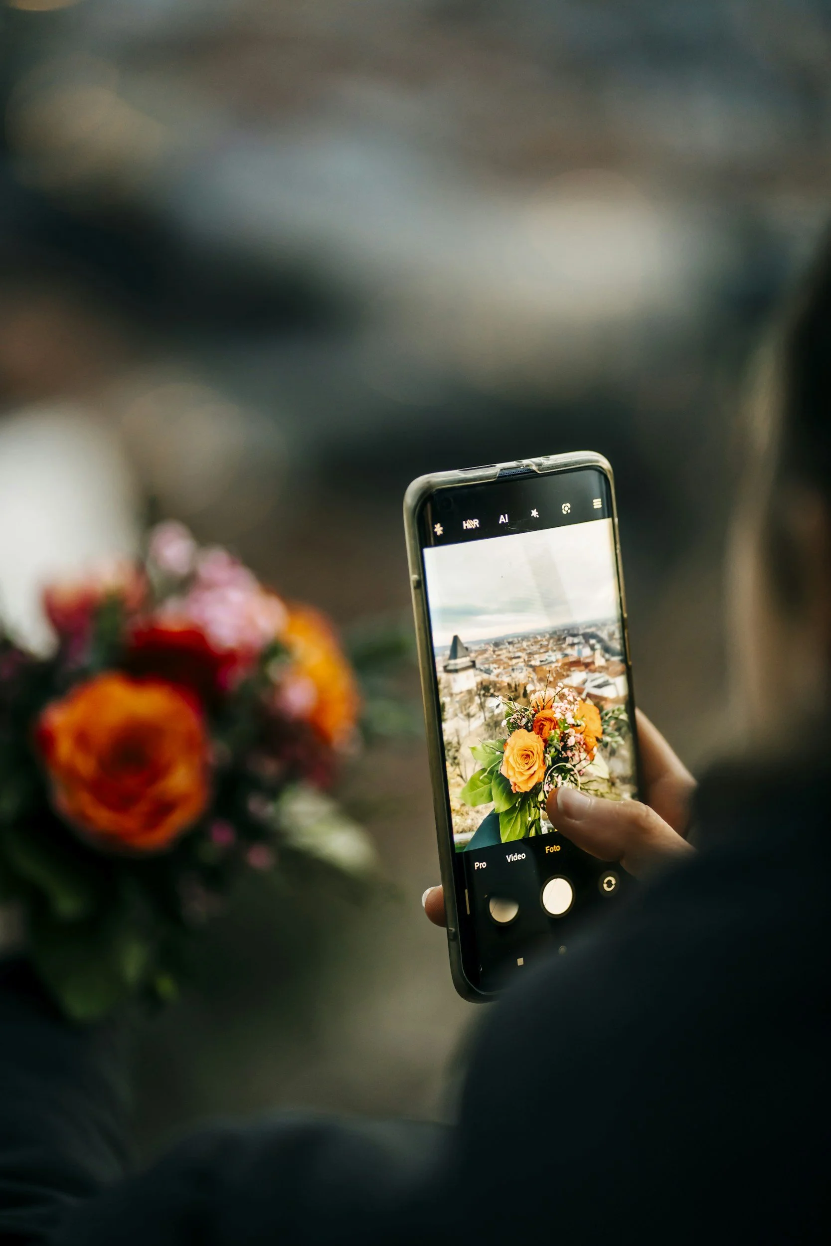 Person taking a photo of a vibrant bouquet of flowers with an orange and pink color scheme on their smartphone. The scene captures the flowers in focus on the phone's screen, with a blurred background.
