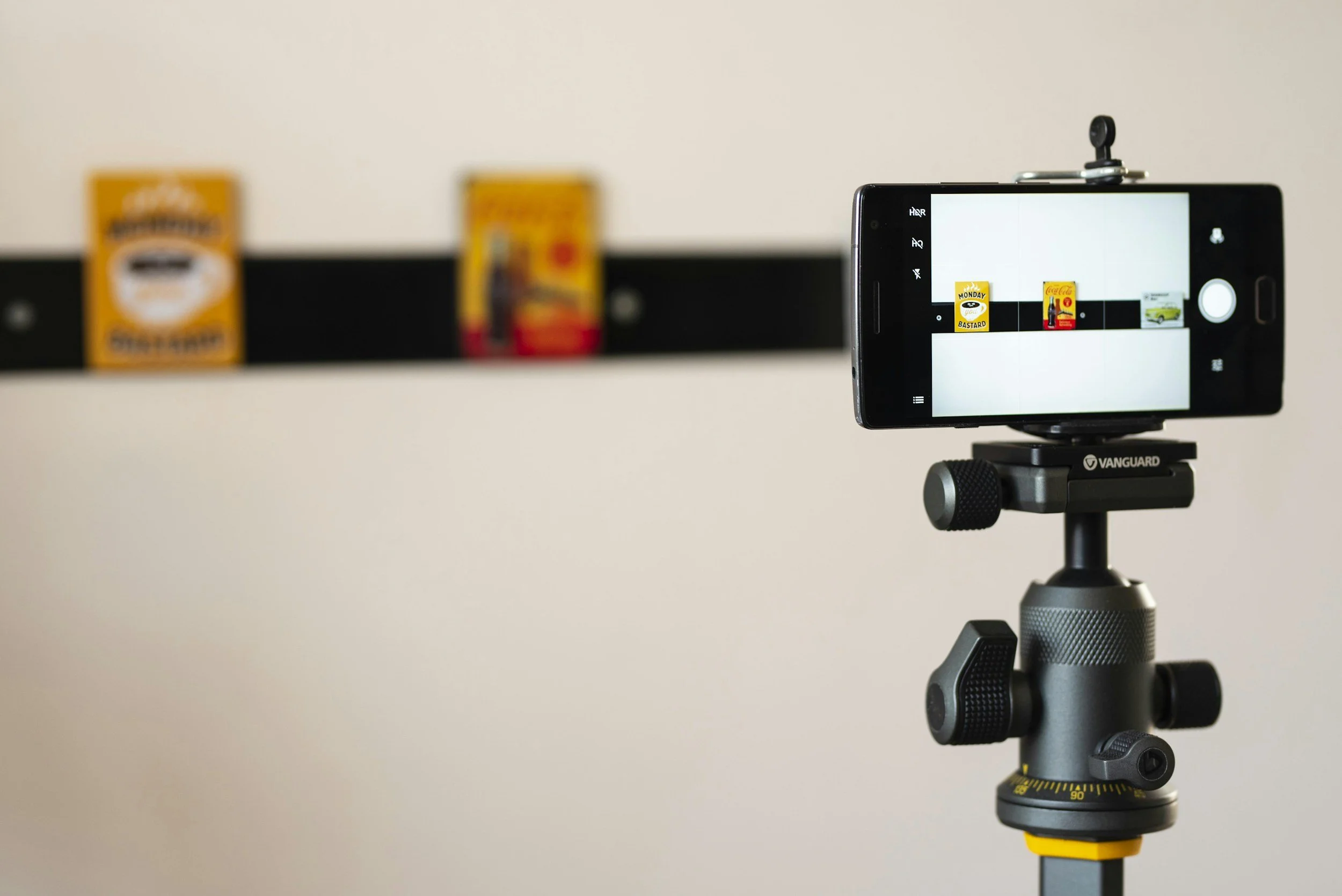 A smartphone mounted on a tripod, taking a photo of three small boxes on a black shelf against a plain wall.
