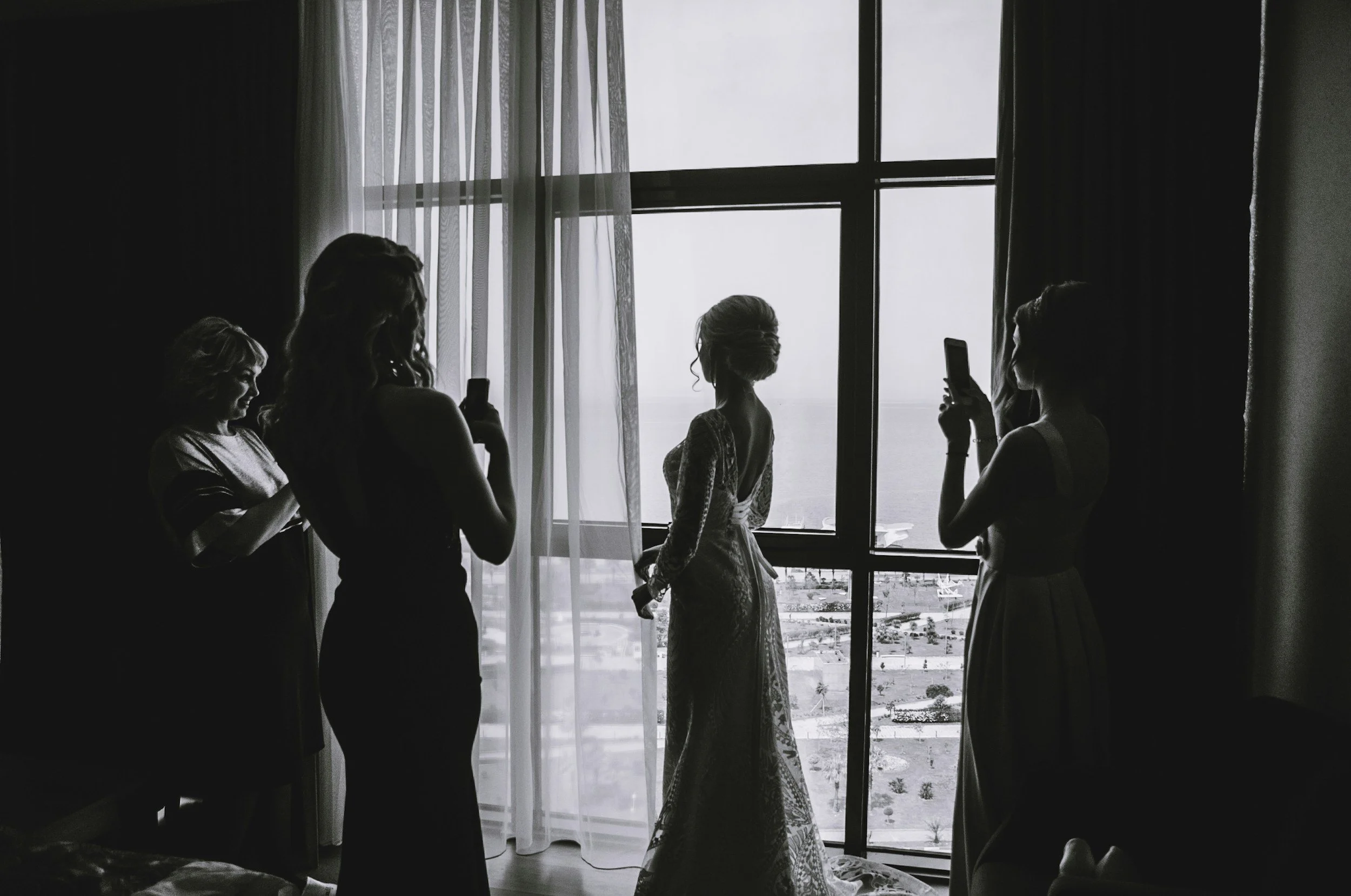 Silhouettes of five women, one in a wedding dress, standing by a large window with sheer curtains, using their phones, in a dimly lit room.