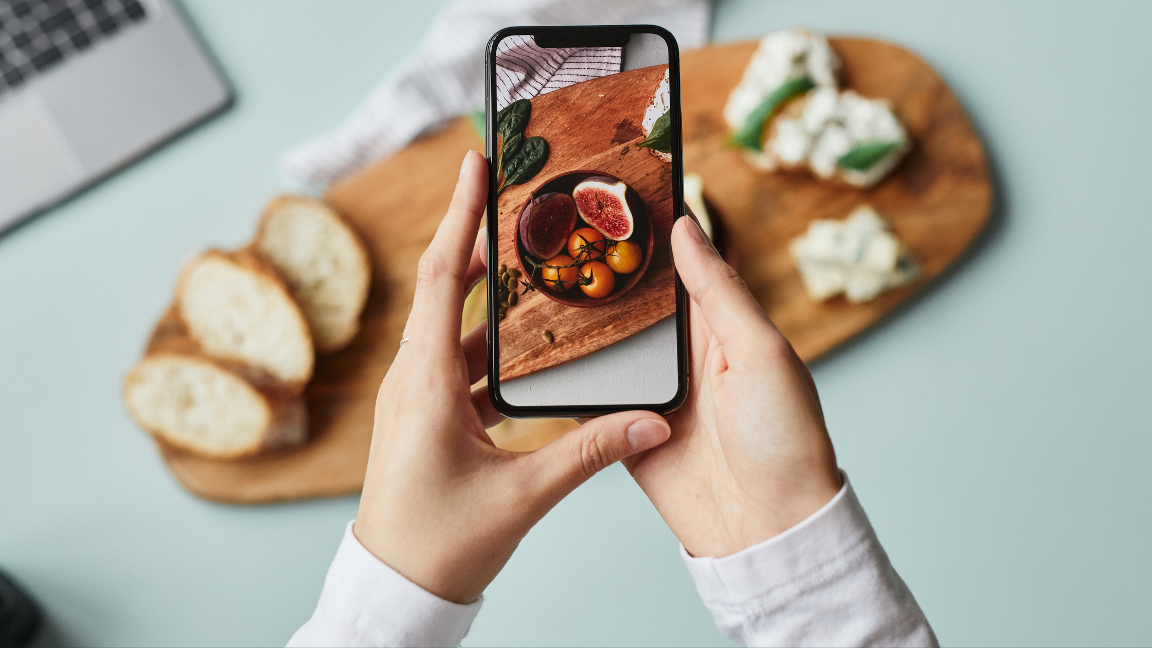 Person taking a photo of a wooden cutting board with a bowl of assorted fruit, including figs and cherry tomatoes, with slices of bread and cheese in the background.