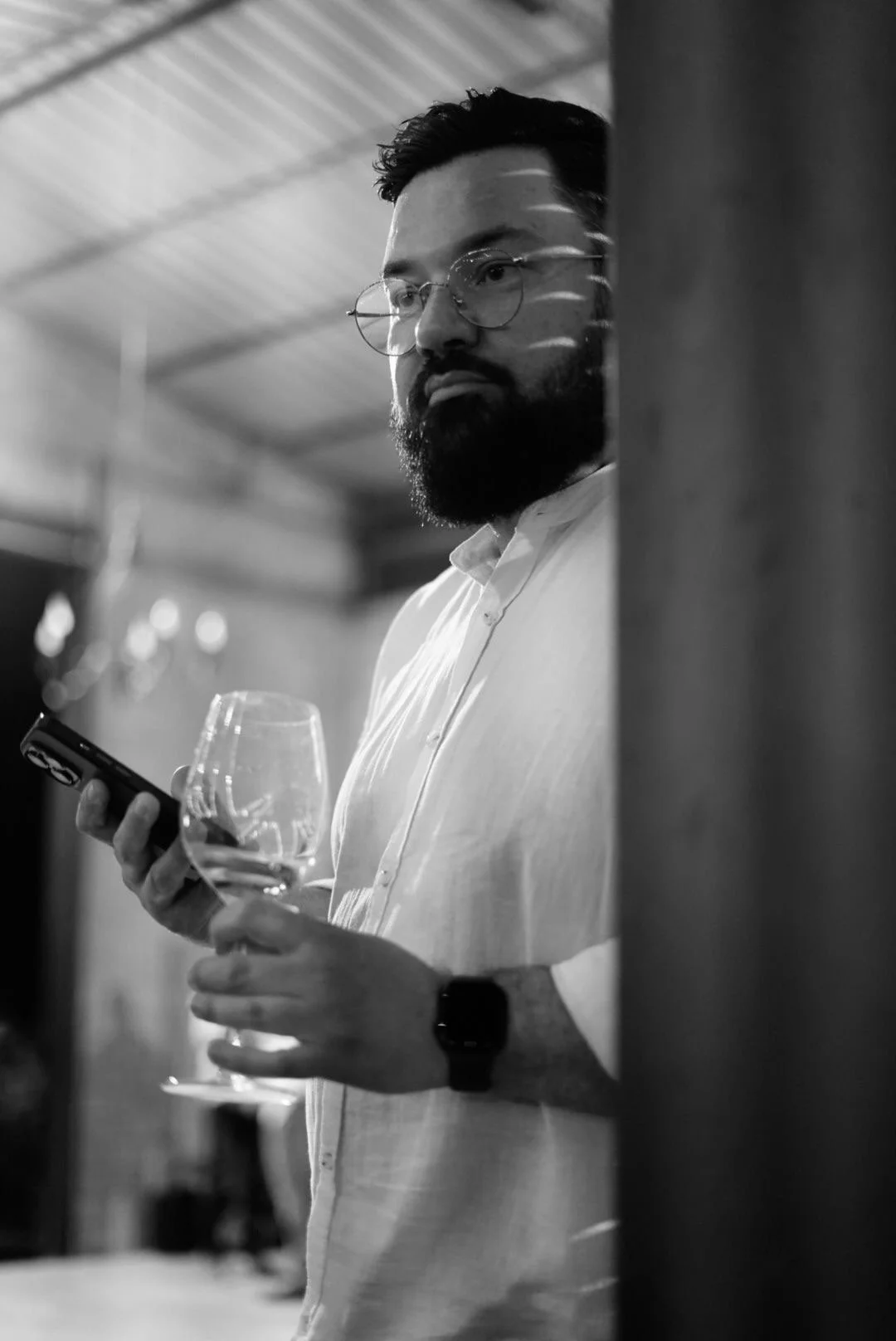 A bearded man with glasses in a white shirt holding a wine glass and a smartphone, standing near a wall, in a black-and-white photo.