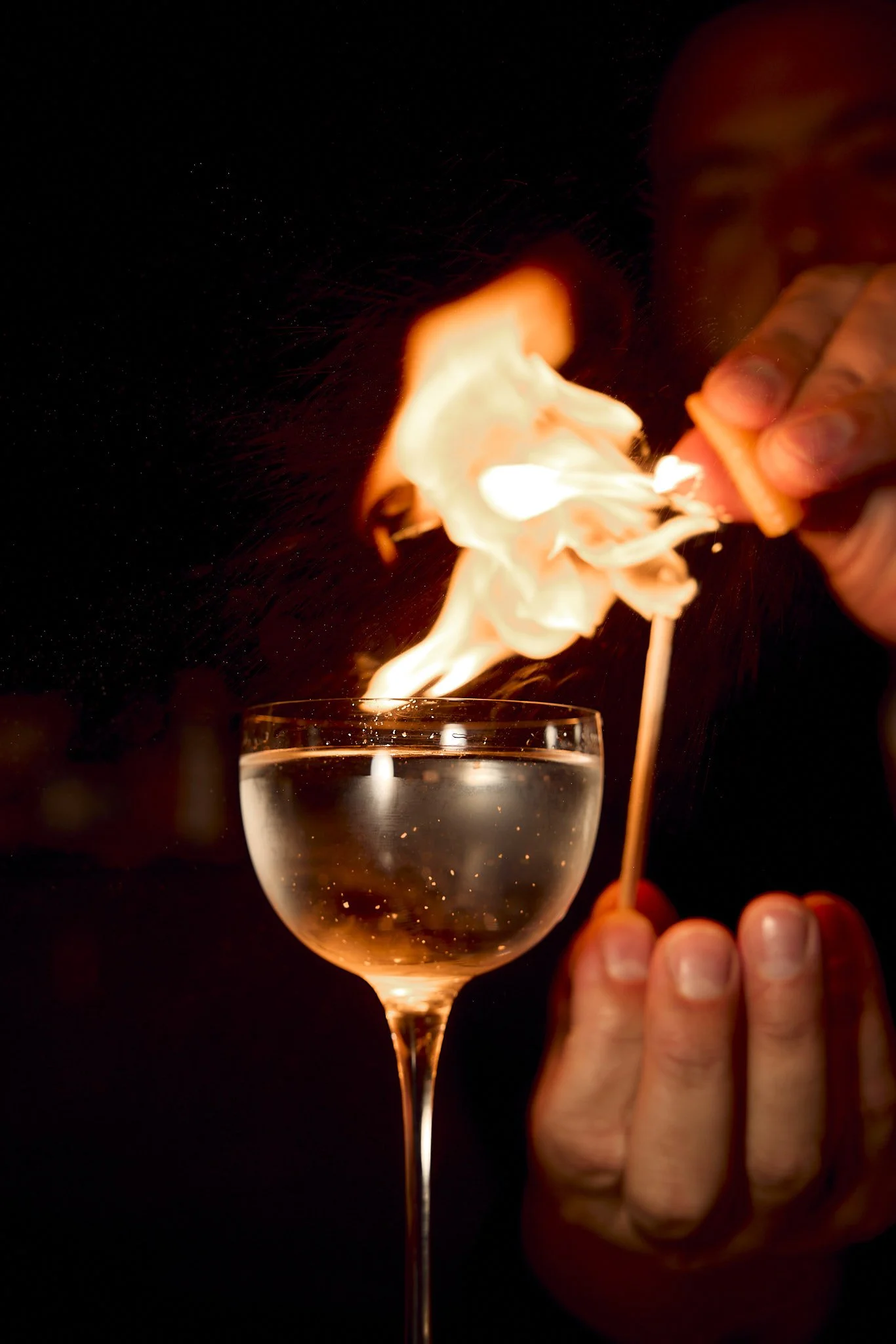 A person holds a matchstick with a lit match, igniting a flame in a glass of water, creating a dramatic visual effect.