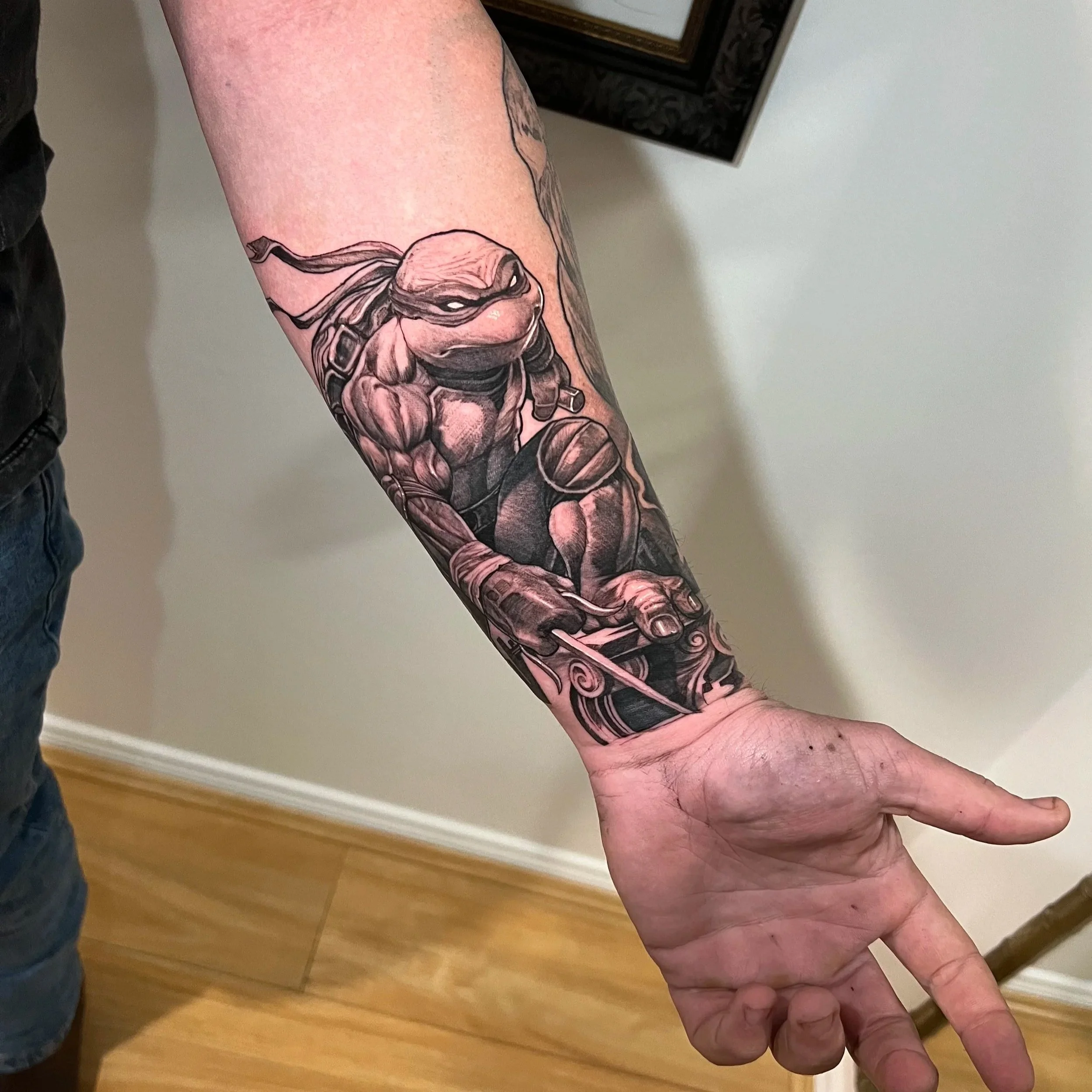 Close-up of a person's arm with a colorful tattoo of a robot or mannequin resembling a human figure with mechanical parts, set against a plain indoor background.