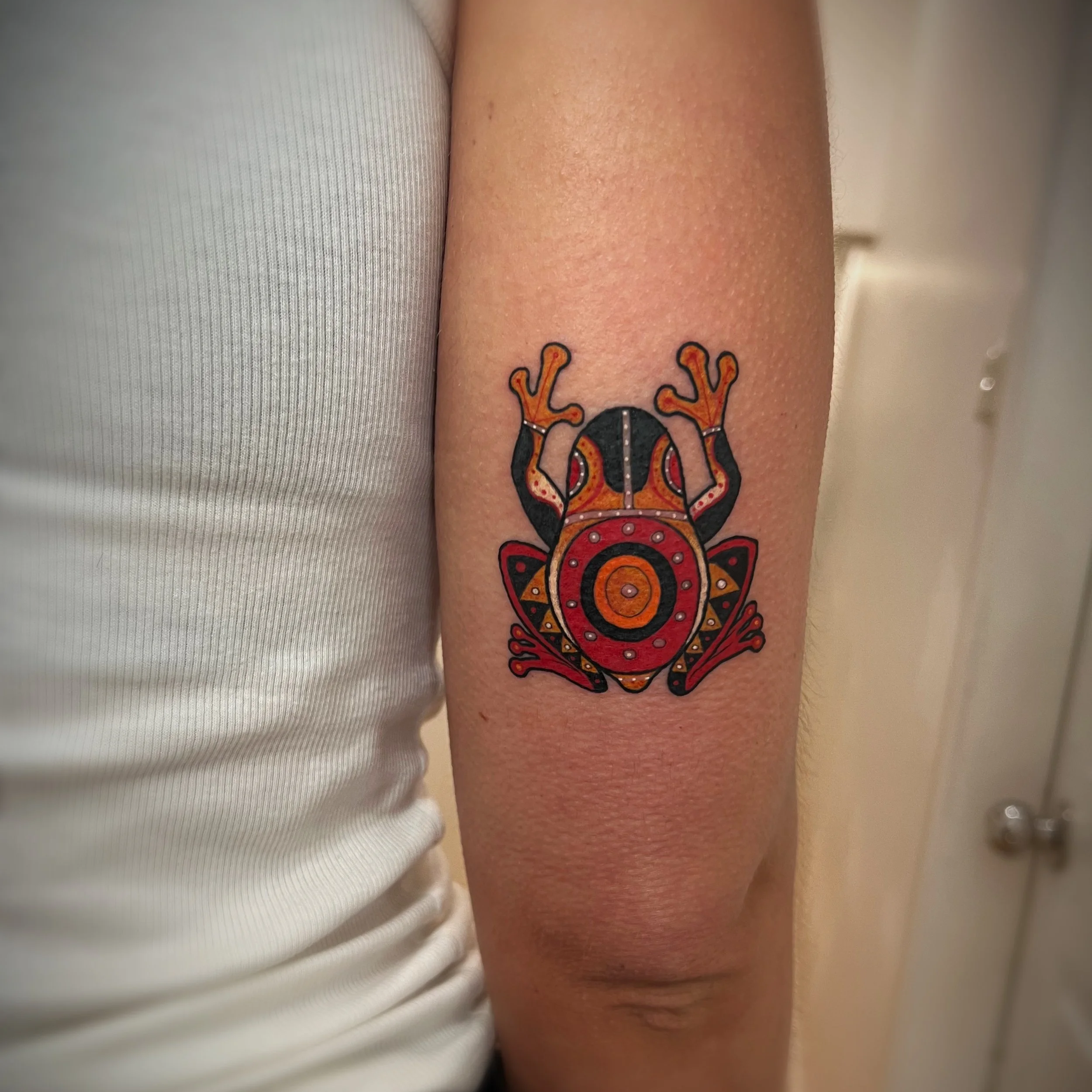 Close-up of a person's arm with a colorful bug-themed tattoo near the wrist, wearing a white short-sleeve shirt.