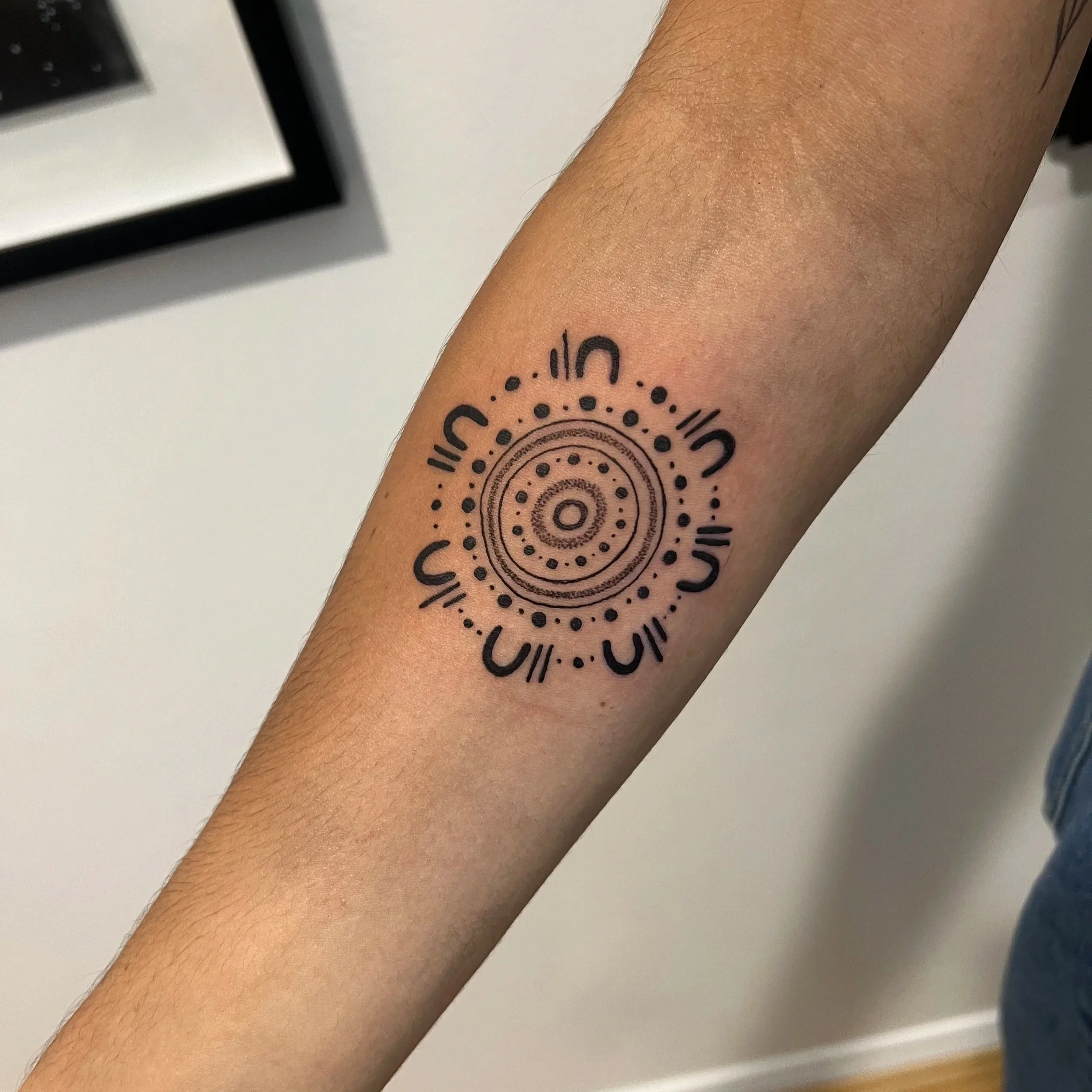 Close-up of a person's arm with a tattoo featuring concentric circles and dotted patterns, the arm is resting on a flat surface, with part of a black object and a white wall visible in the background.