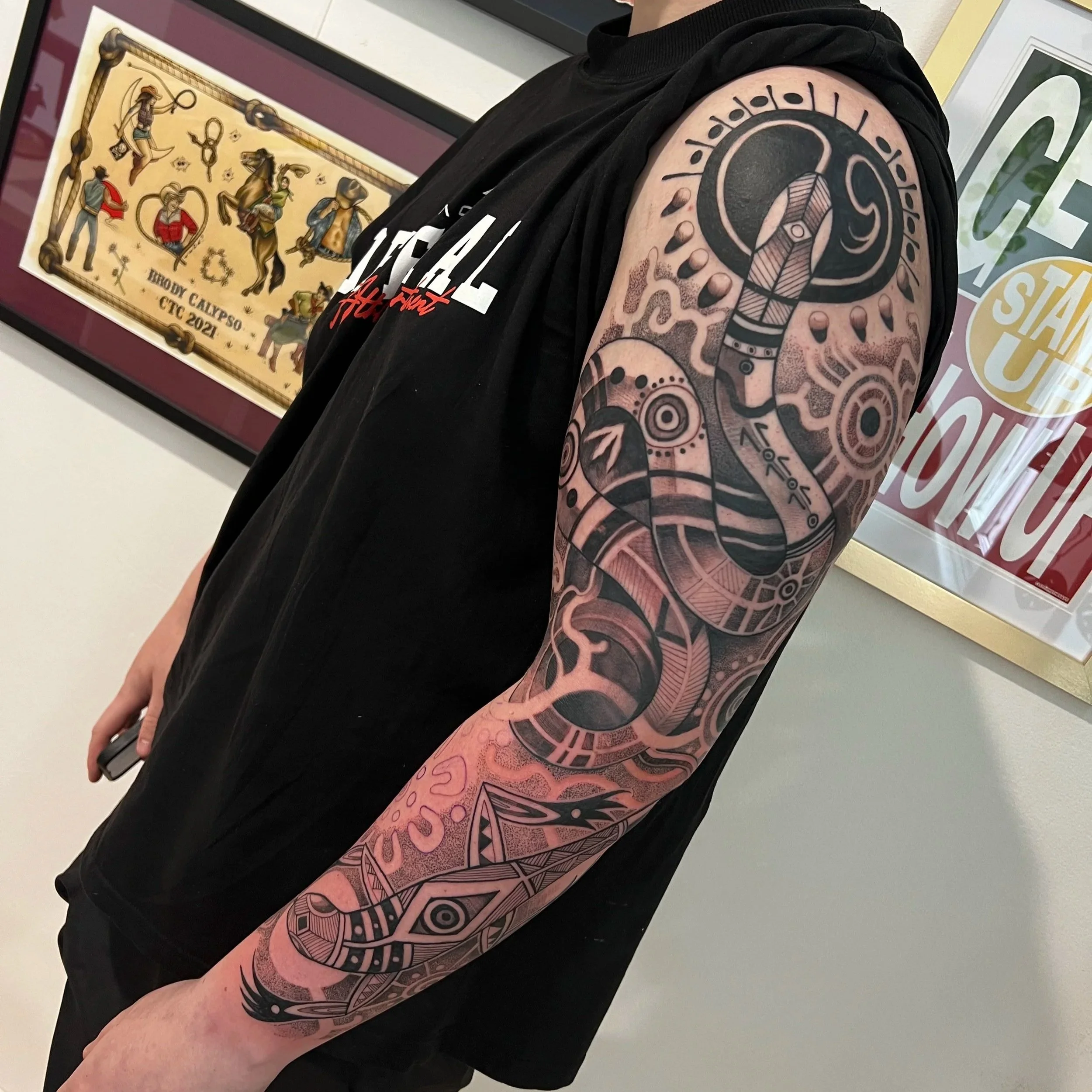A person's arm with a large black and grey Polynesian style tattoo, standing next to framed artwork featuring various figures, including a mermaid and a dog, on a beige background.