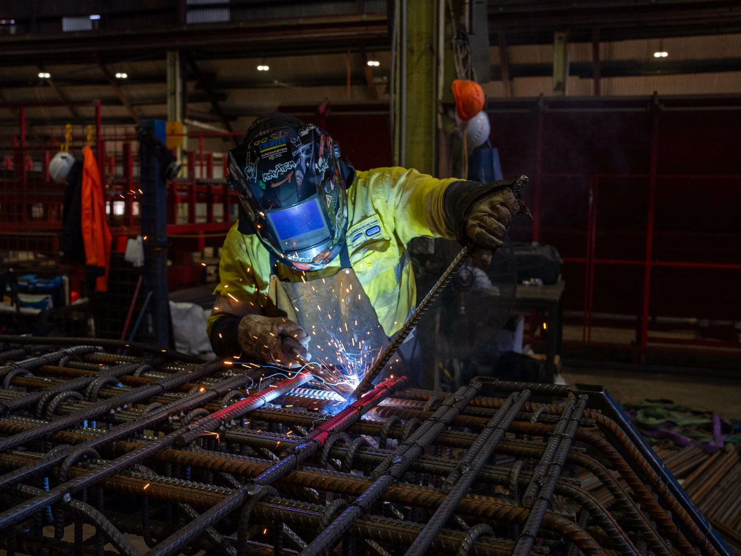 Welder in hi-vis safety gear assembling reinforcement cage inside Carrabay workshop.