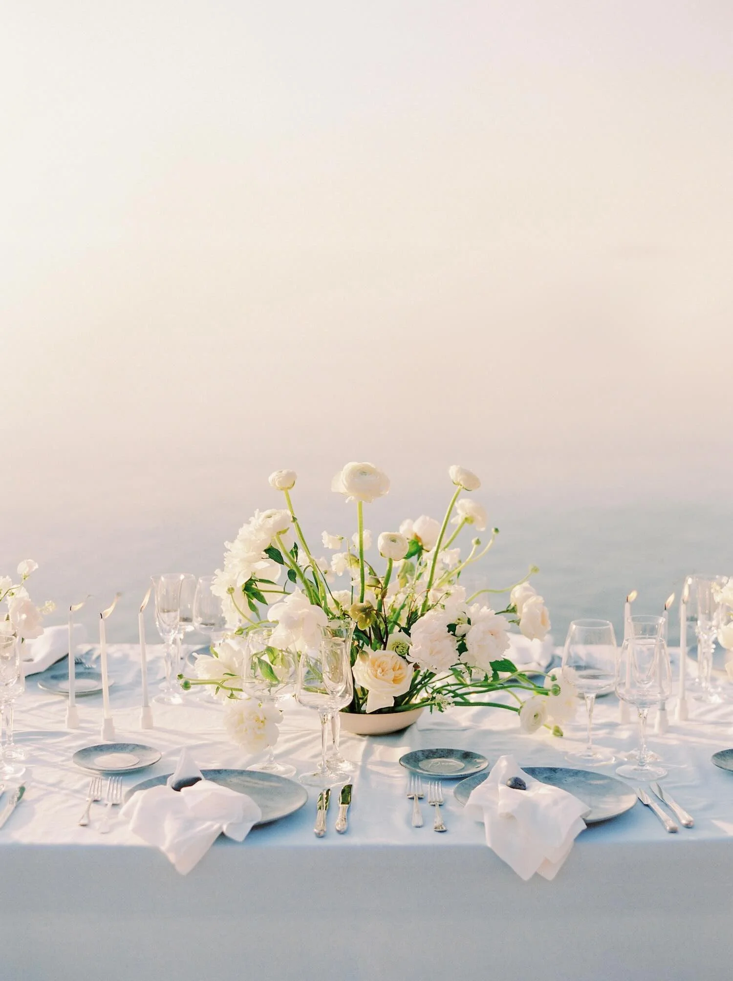 Elegant outdoor dining table with white floral centerpiece, glassware, plates, and napkins, set against a coastal background.