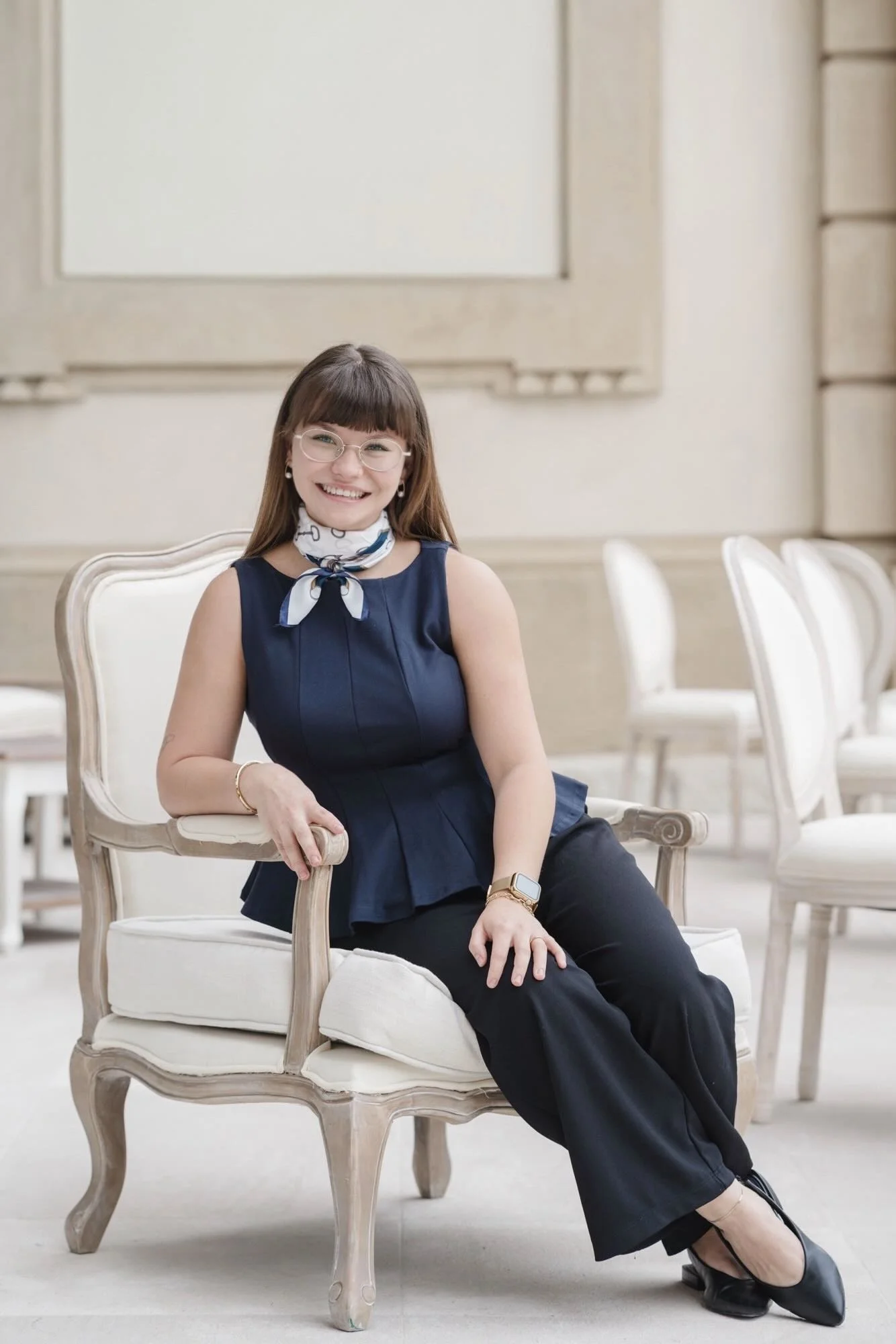 A woman with long brown hair, glasses, and a white scarf with blue design, smiling and sitting on a cream-colored vintage-style chair in an elegant room. She is wearing a navy sleeveless top, black pants, and black heels.