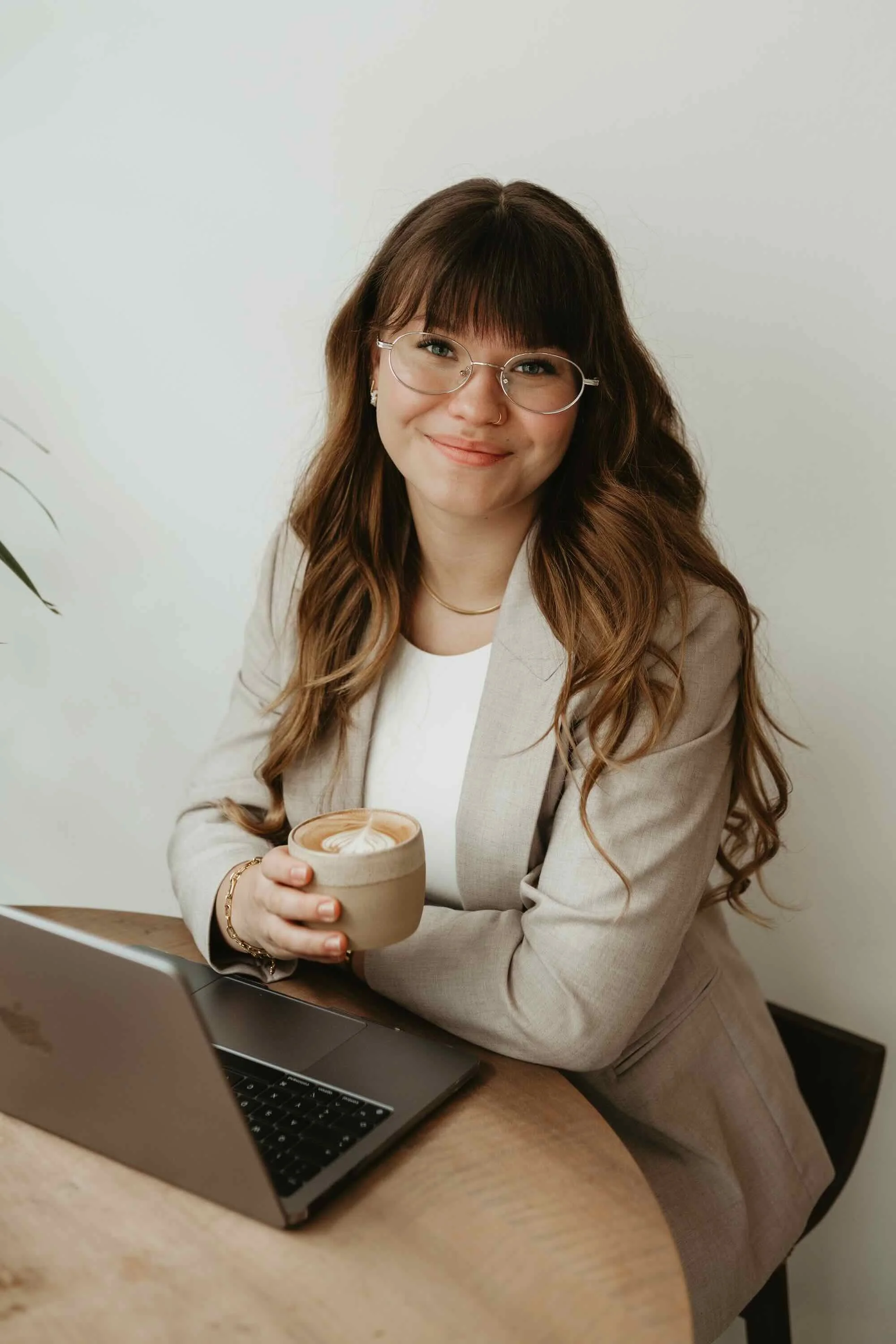 A woman with long wavy hair, glasses, and a beige blazer sitting at a wooden table, holding a cup of coffee or tea, with a laptop in front of her, smiling at the camera.