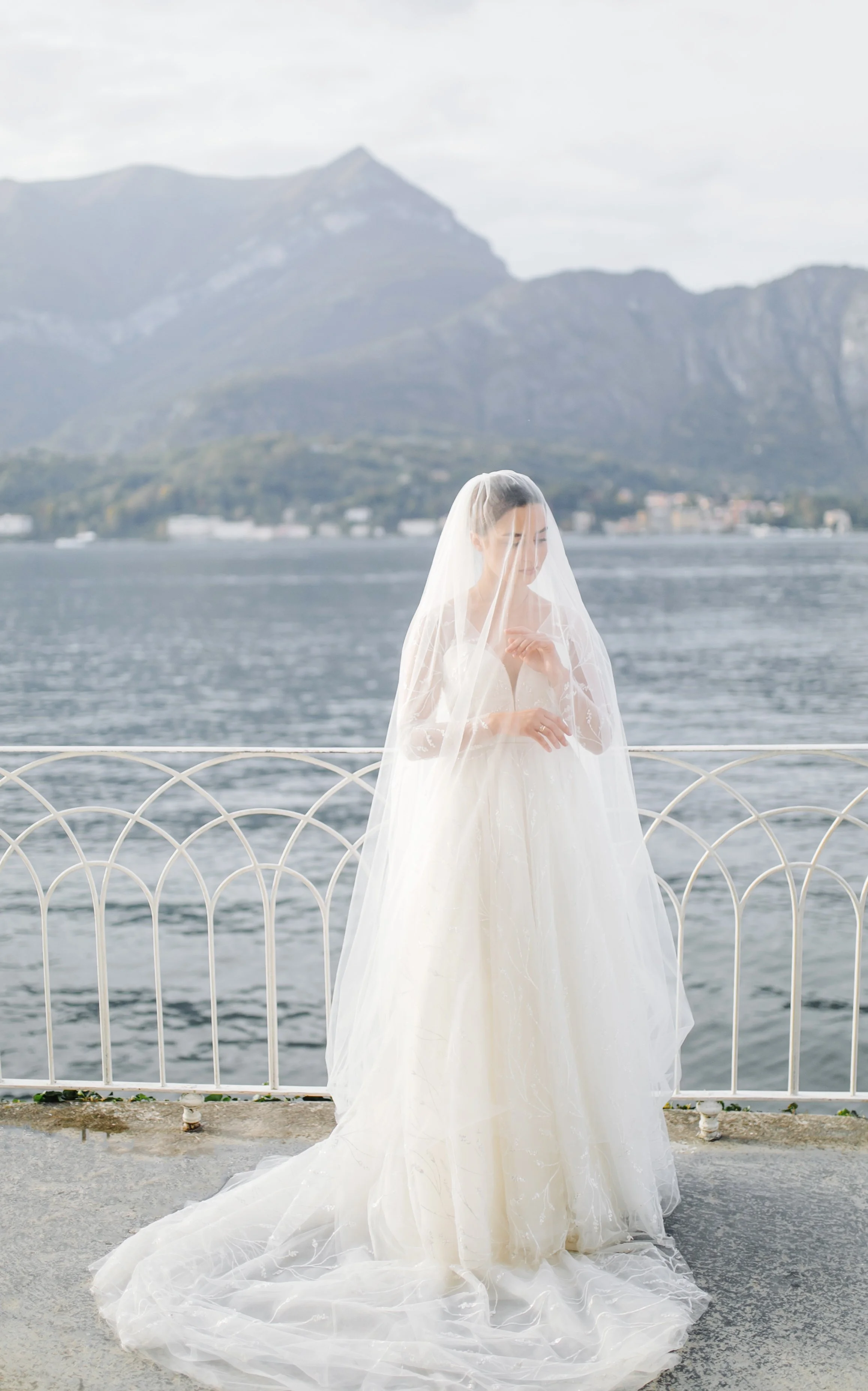 Bride in a white wedding dress and veil standing by a waterfront with mountains in the background.