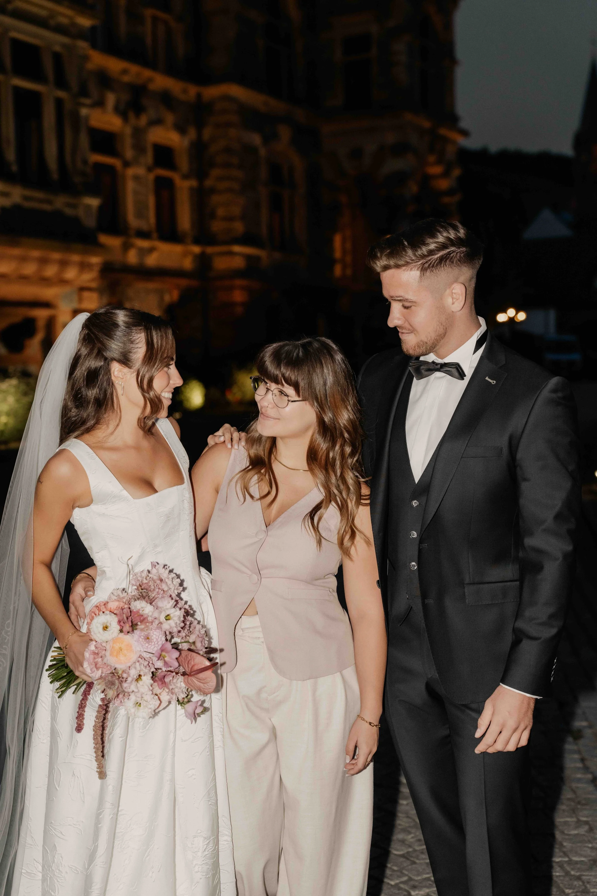 A bride and groom stand with two women outdoors at night, smiling and looking at each other, with a large historic building in the background.