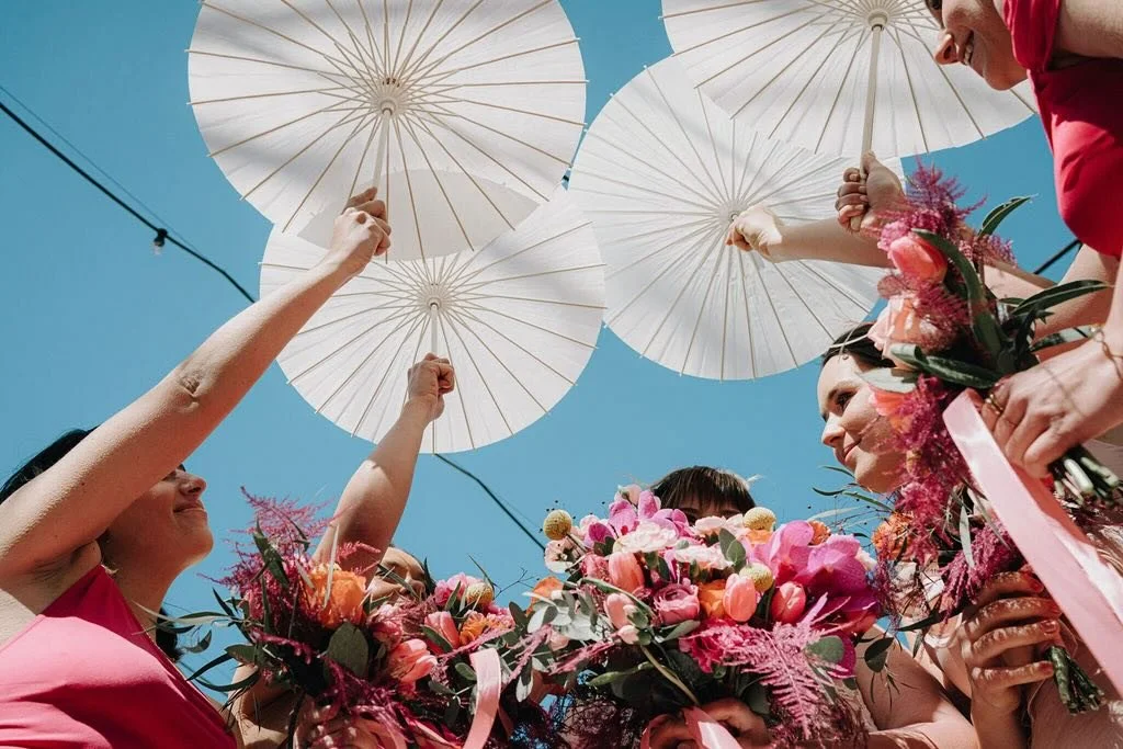 Women holding white parasols overhead at a celebration, with pink and purple floral bouquets, under a clear blue sky.