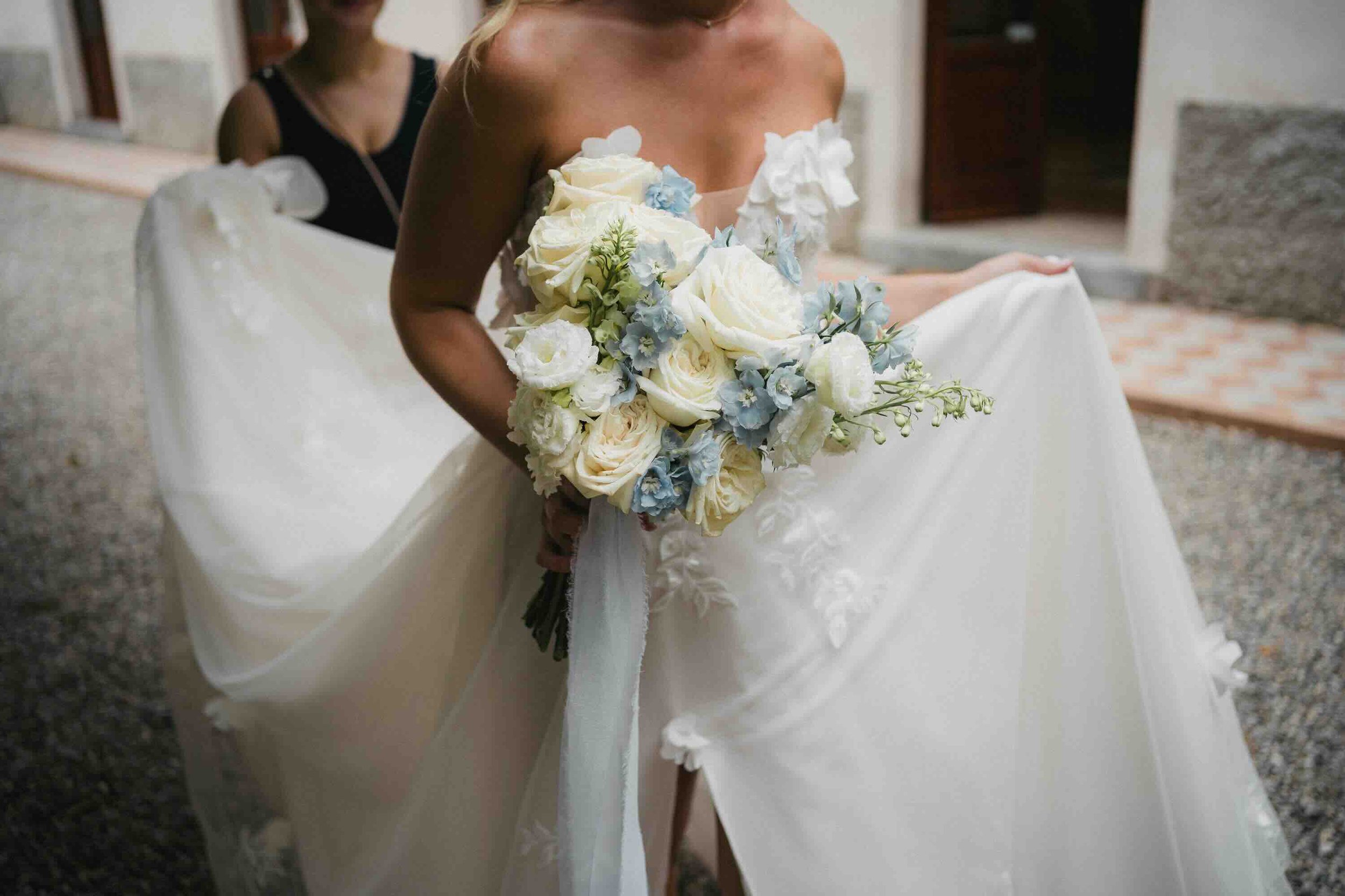 A bride holding a bouquet of white and pale blue flowers, wearing a white wedding dress with floral details, outdoors near a building.