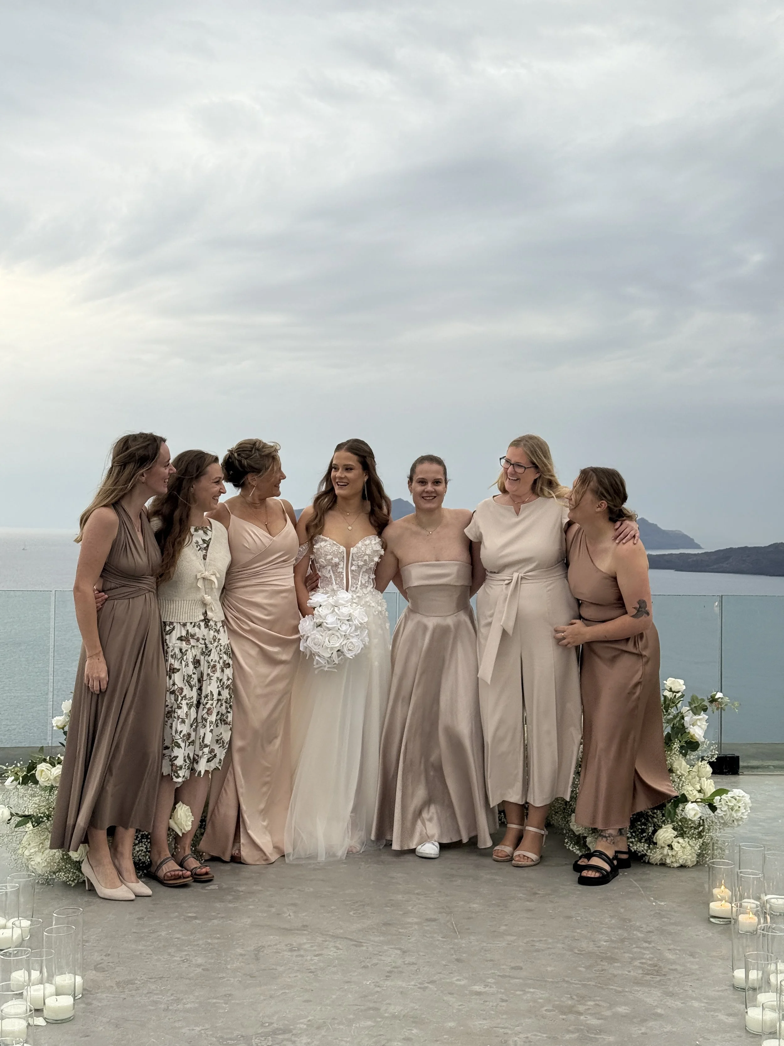 Group of women at a wedding, standing on a terrace with an ocean view, dressed in formal dresses, with floral decorations and candles.