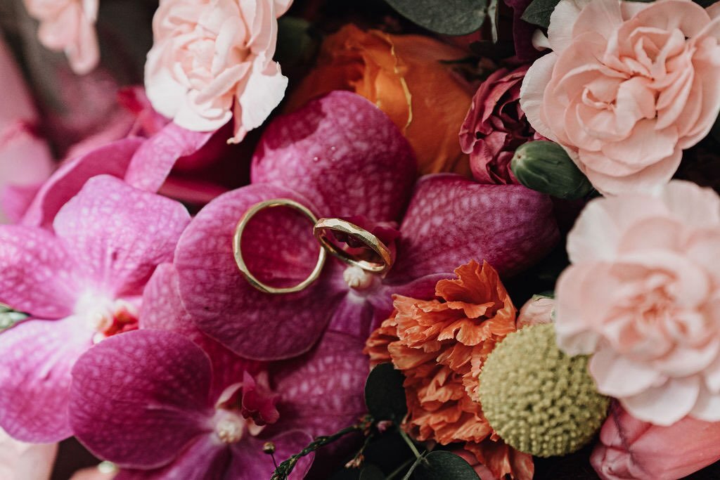 Two gold wedding rings resting on pink orchid flowers surrounded by various pink and orange flowers.