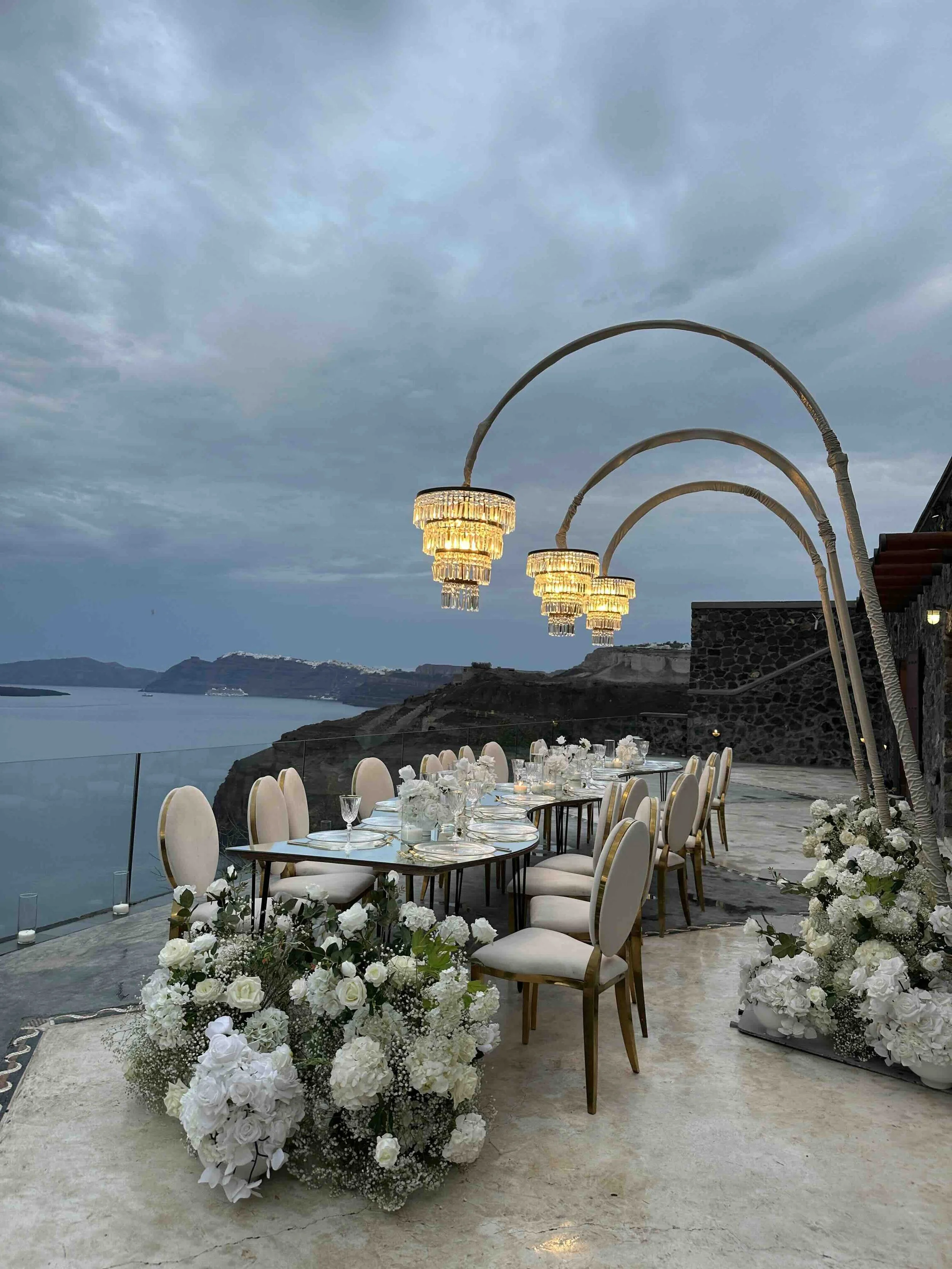 Elegant outdoor dining area with a long table decorated with white flowers, surrounded by white chairs, under ornate chandeliers, with a scenic view of a body of water and hills at dusk.