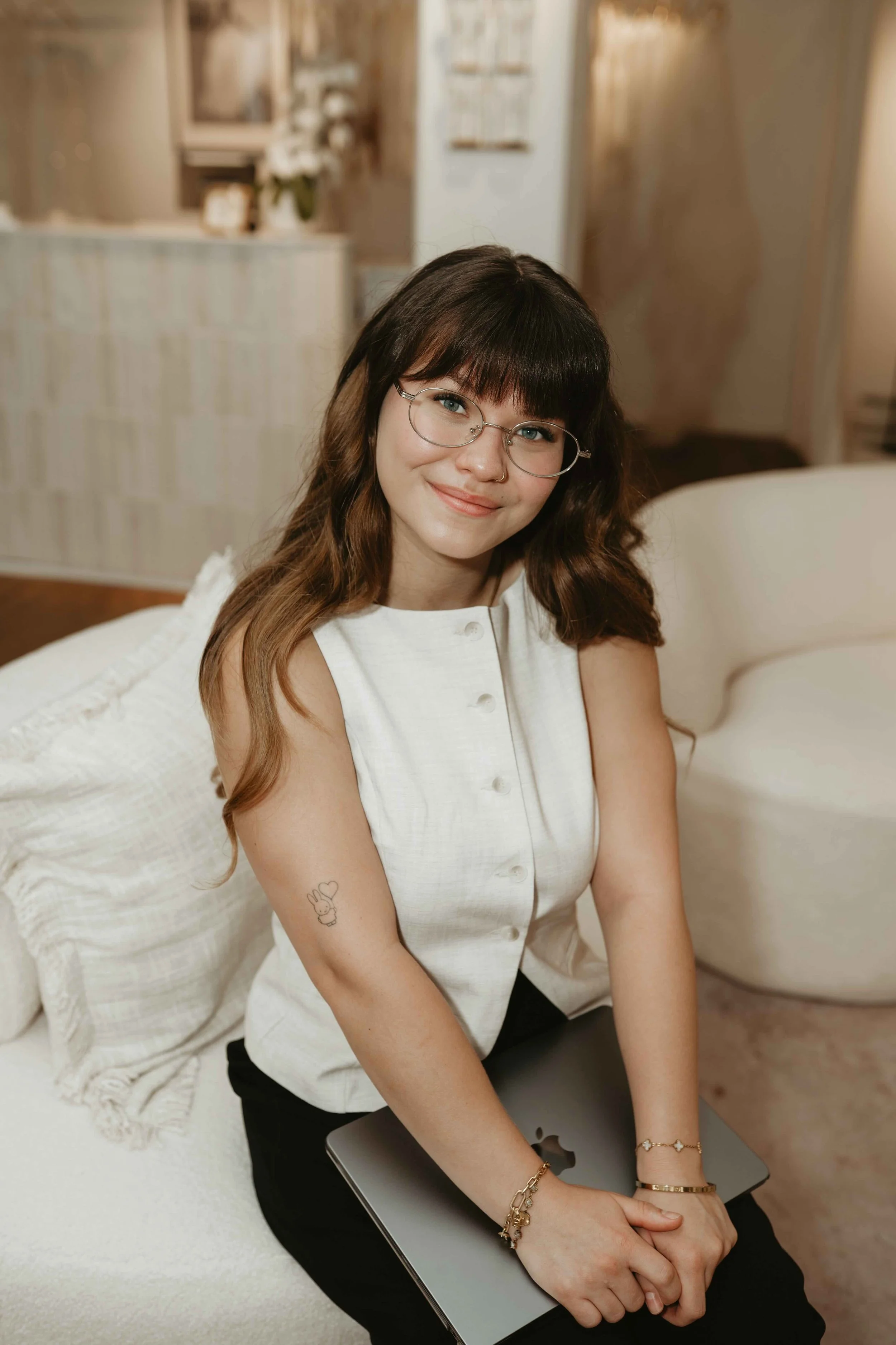 A young woman with glasses and a white sleeveless top sitting on a cream-colored couch in a cozy living room, holding a closed laptop on her lap, smiling softly at the camera.