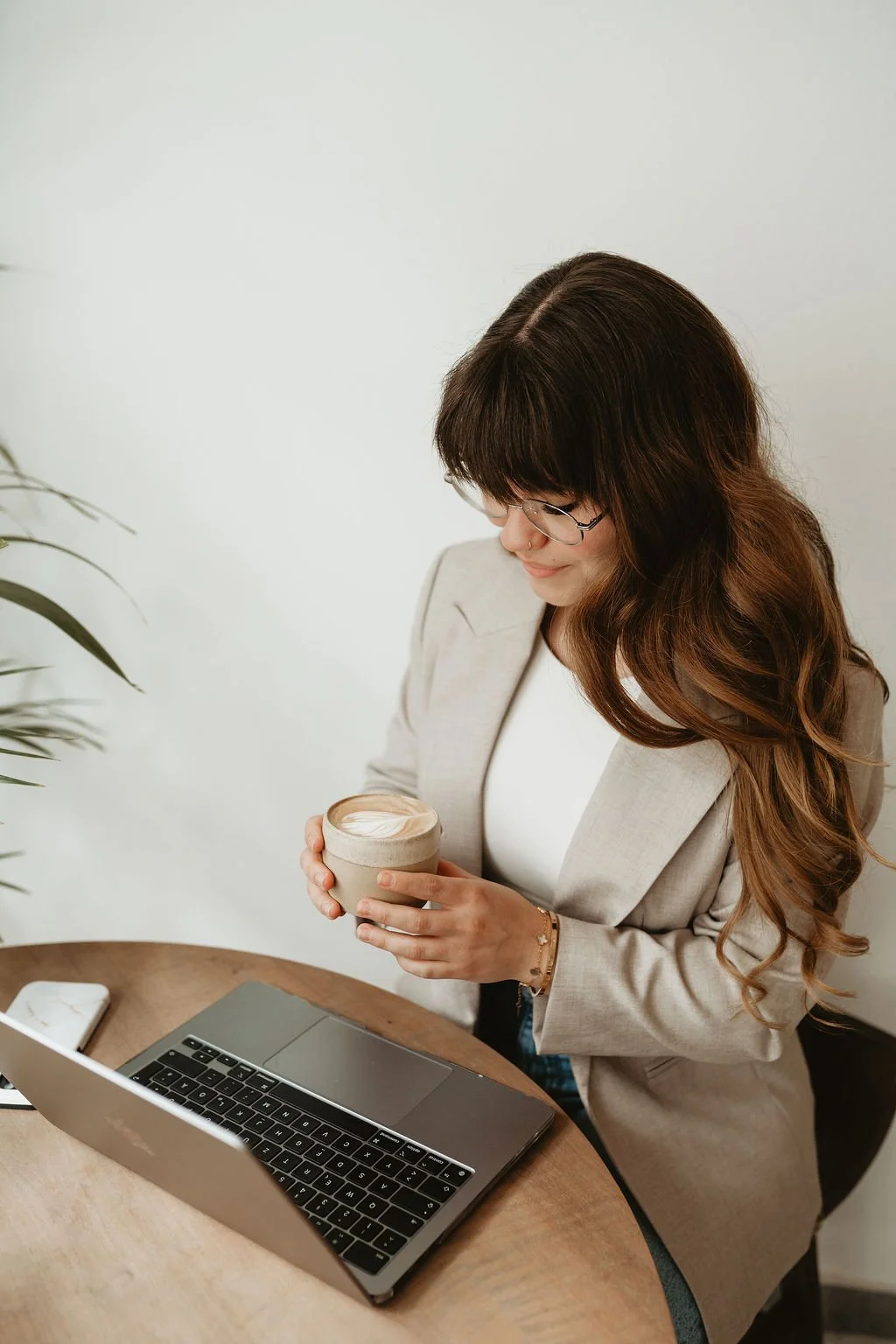 A woman with long brown hair and glasses, wearing a beige blazer and a white shirt, holding a coffee cup while sitting at a wooden table with a laptop and smartphone, inside a bright room with a white wall and a plant.
