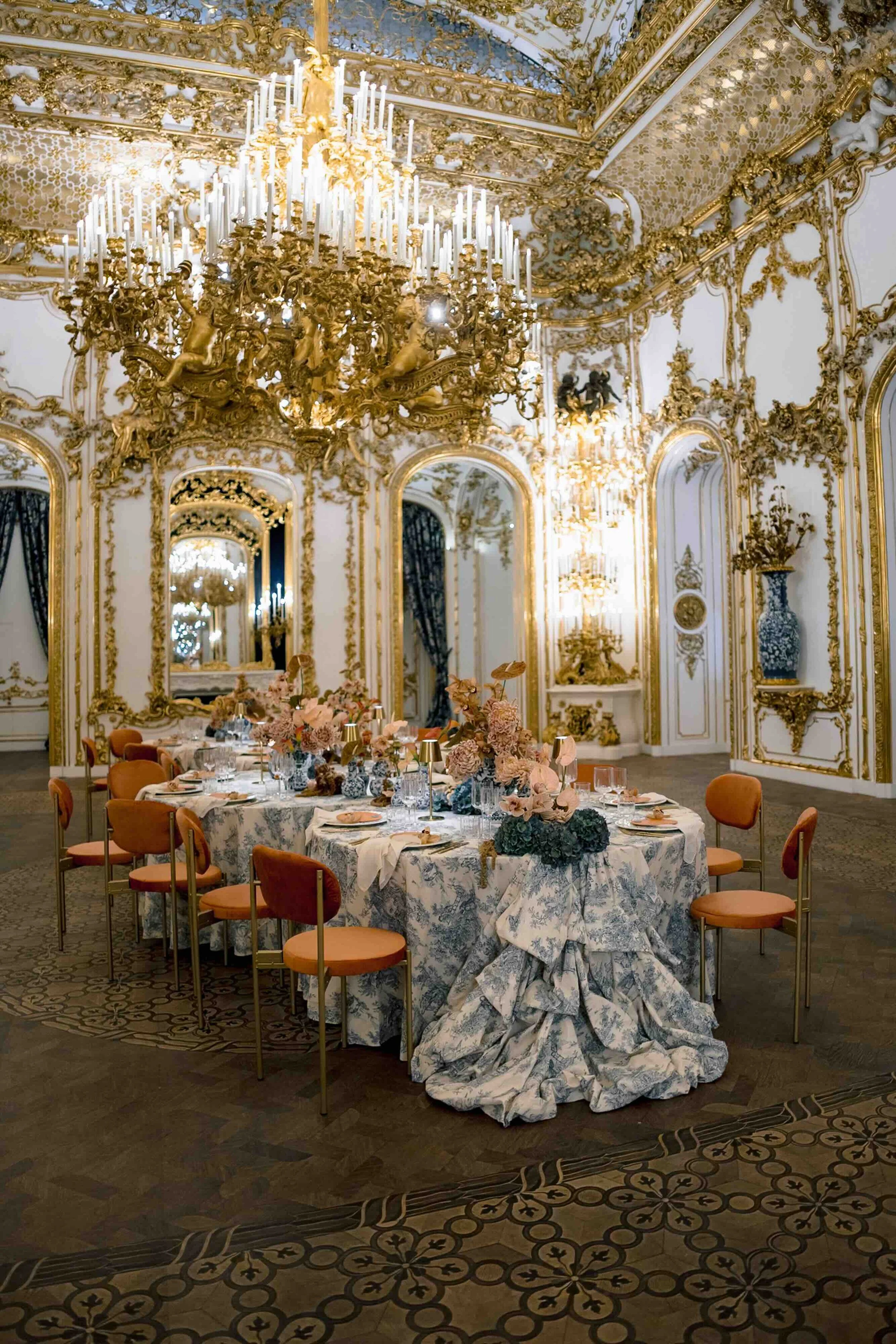 An elegant, ornate banquet hall with gold detailing, a large chandelier hanging from the ceiling, and a long table set for a formal event with floral centerpieces and tableware.