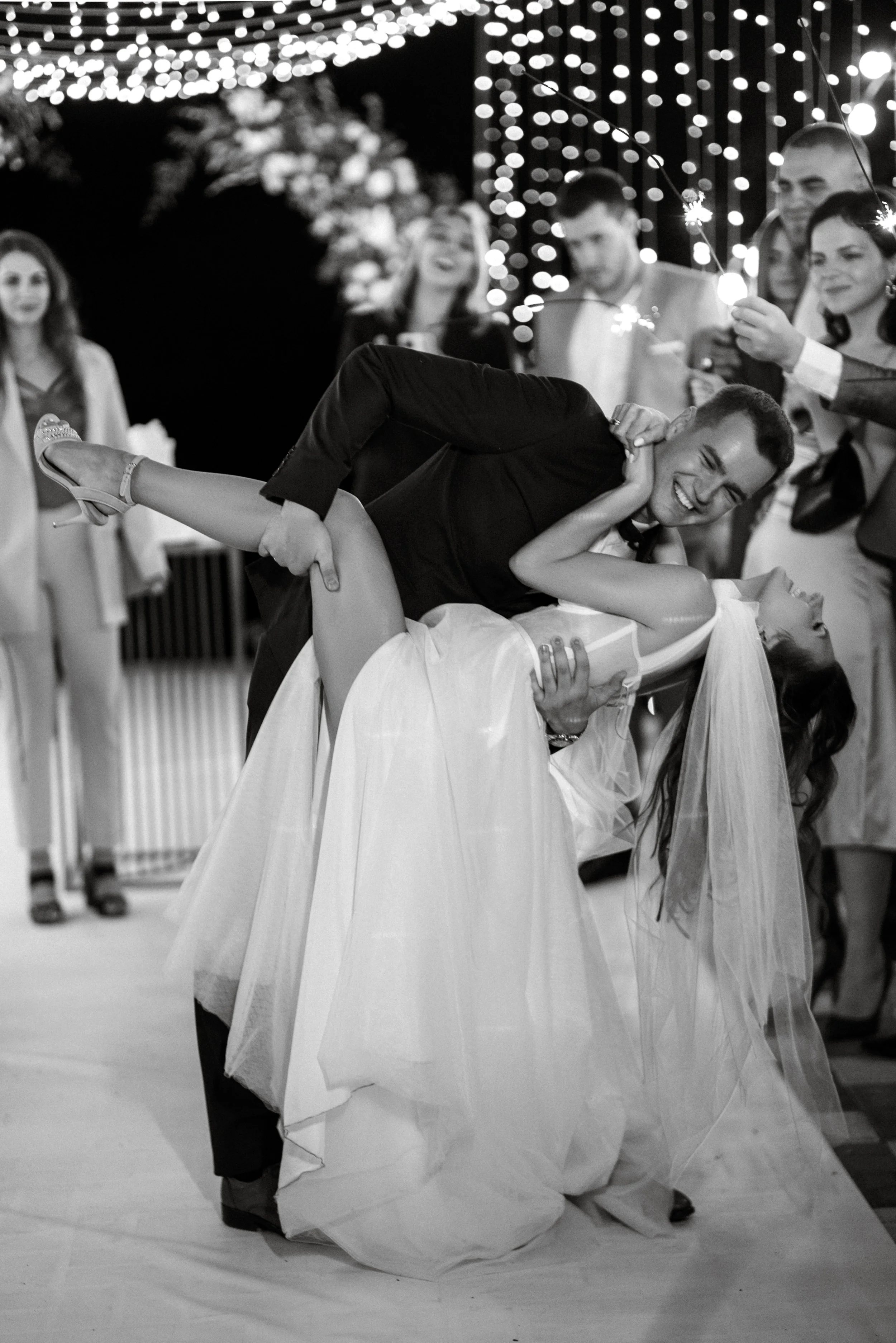 A groom dips and kisses a bride during the wedding dance, surrounded by guests holding sparklers, with string lights overhead at night.