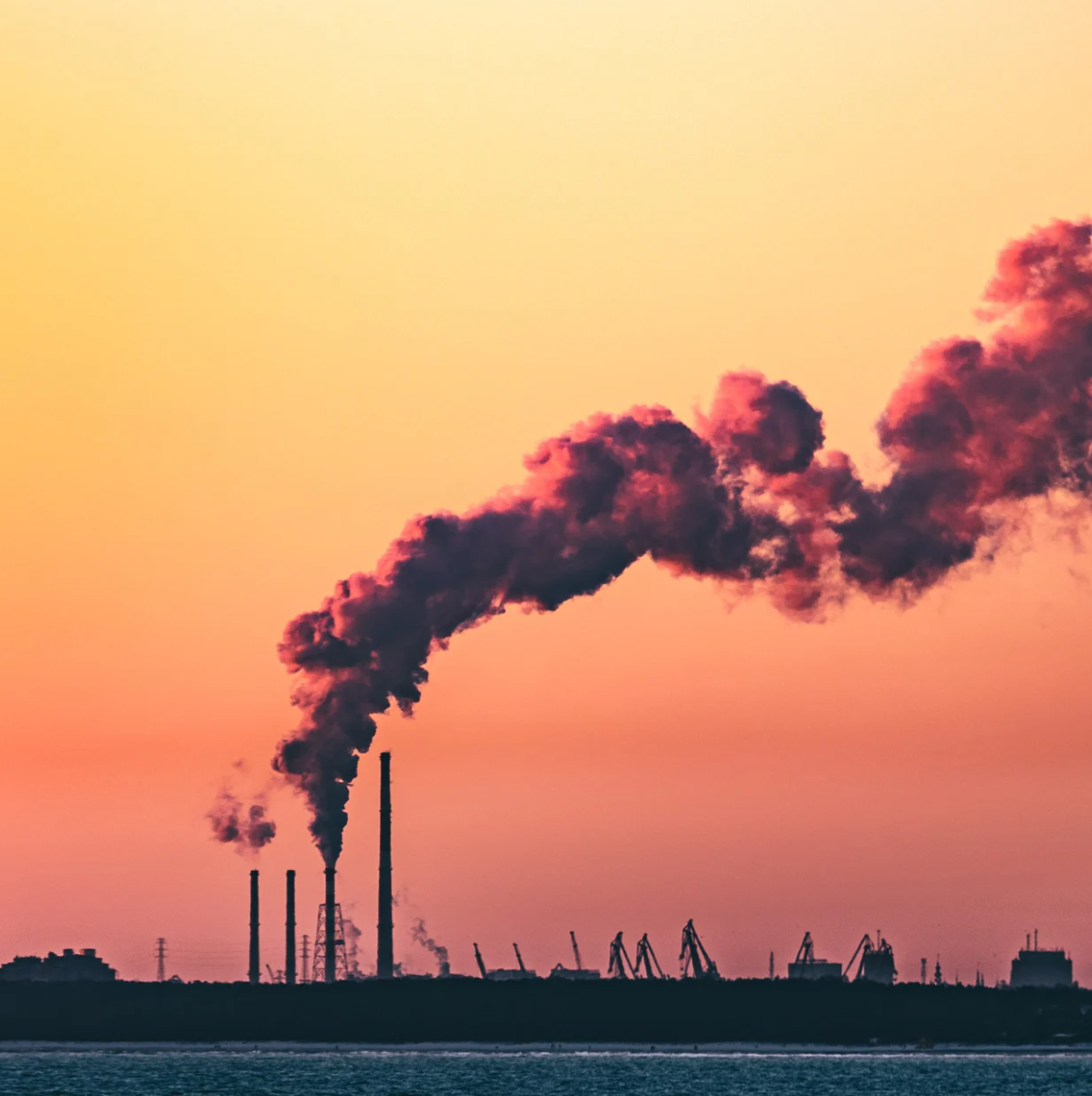 Industrial landscape with smoke rising from multiple smokestacks against a pink and orange sunset sky.