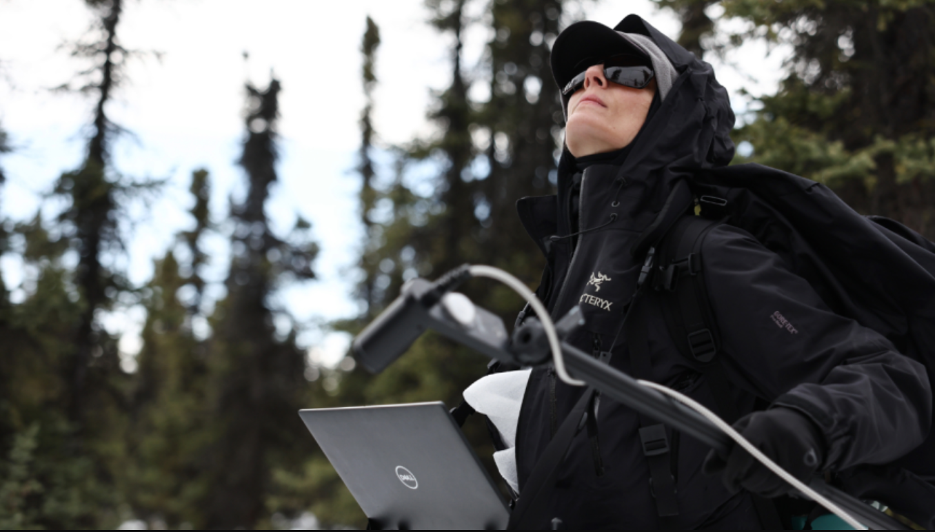 Person outdoors in winter gear, wearing sunglasses, holding a laptop, with trees in the background.