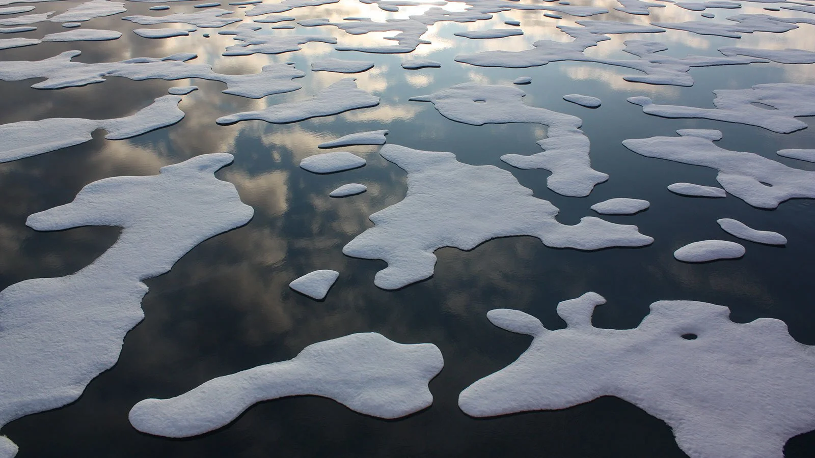 Ice patches floating on a body of water, reflecting the cloudy sky above.