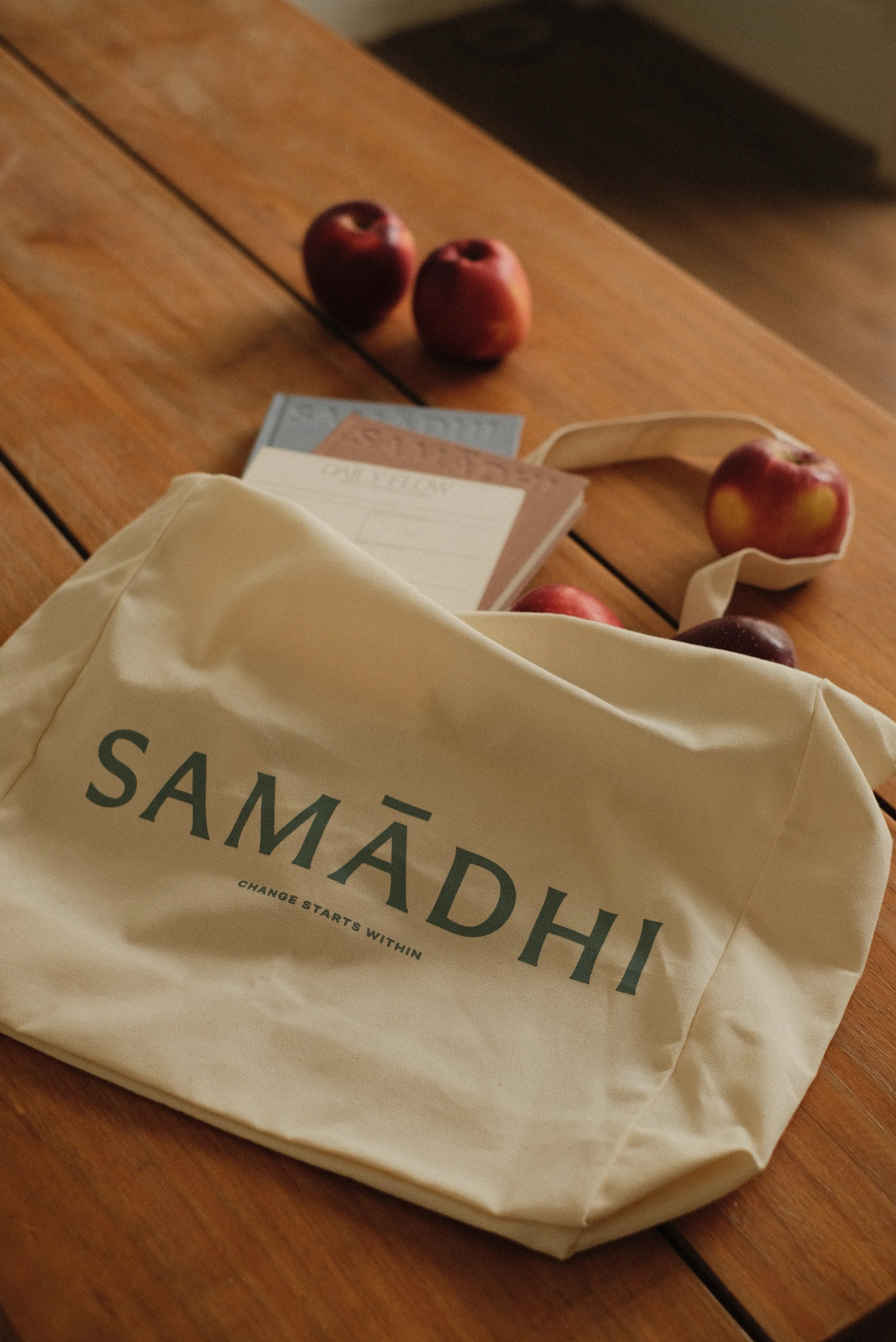 A beige tote bag with the words 'SAMĀDHI' and 'CHANGE STARTS WITHIN' on a wooden table, surrounded by apples, notebooks and reflection journals, including a gratitude journal.