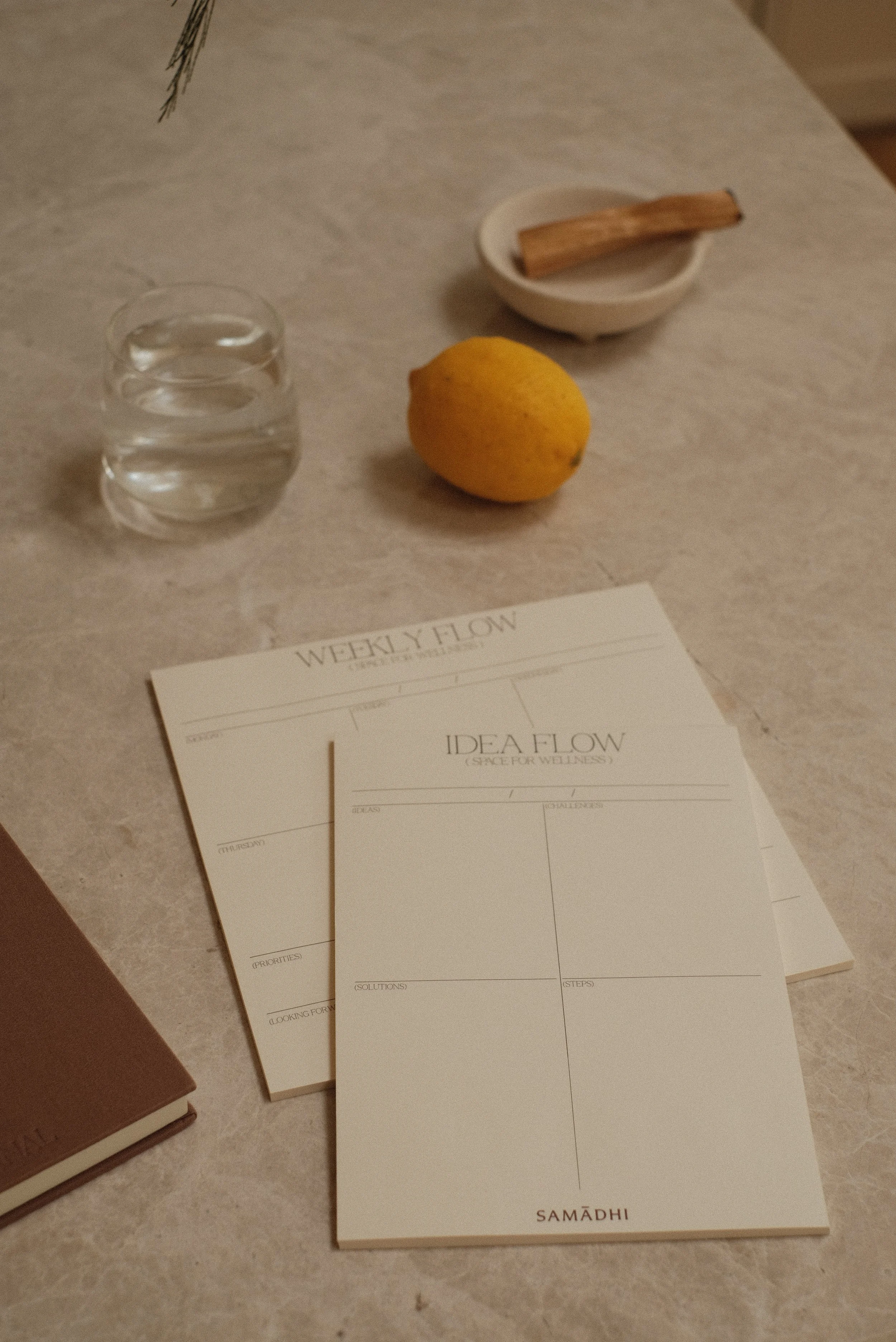 A glass of water, a lemon, a small dish with a cinnamon stick, and wellness planner and idea pads on a marble surface. There is also a brown open reflection journal on the side.