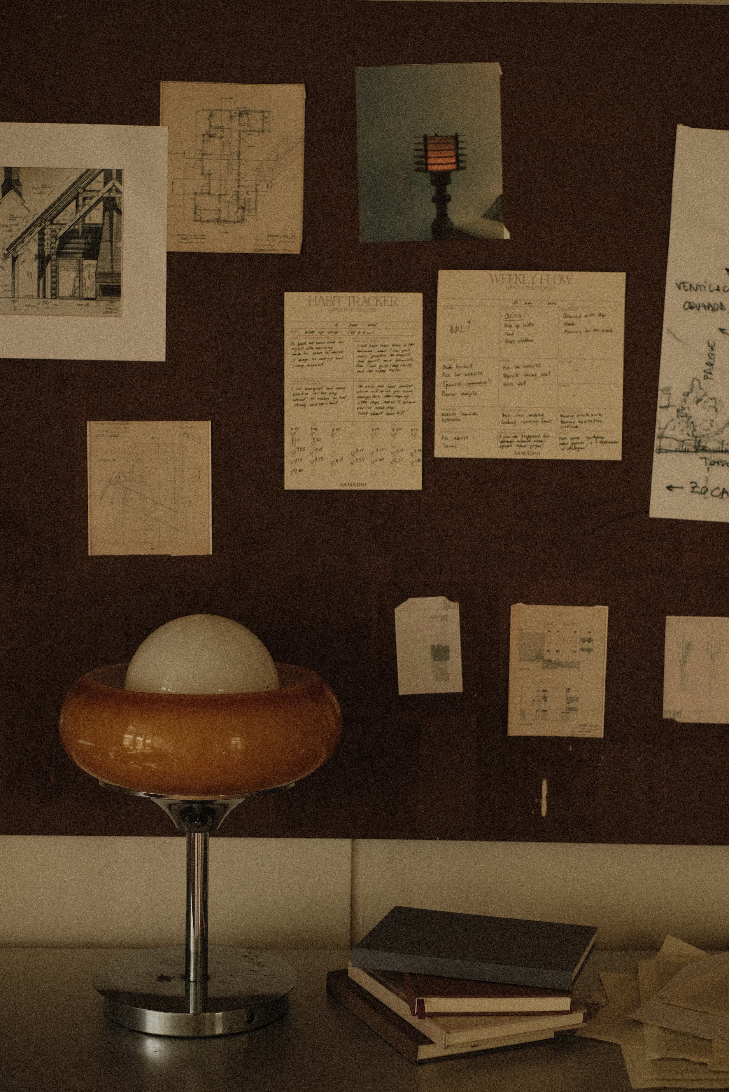 A wooden bulletin board with planner pads and pictures pinned on it. A desk lamp with a globe-shaped light sits on a desk below, alongside a stack of different reflection journals and a gratitude journal.