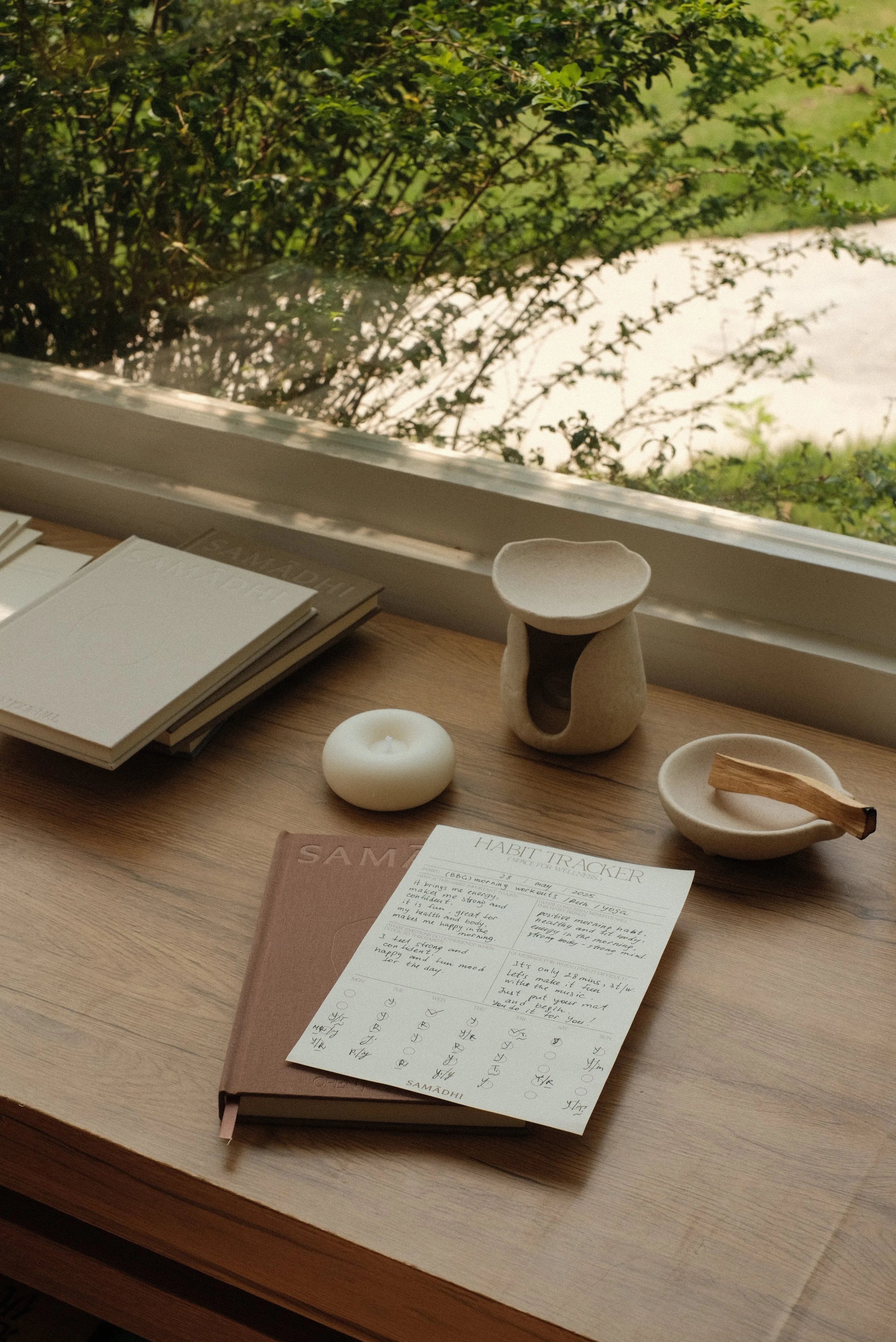 A wooden desk near a window with reflection and gratitude journals, a habit tracker sheet, a white candle, a small ceramic sculpture, a shallow dish with a wooden stick, and a plain white plate, overlooking greenery outside.