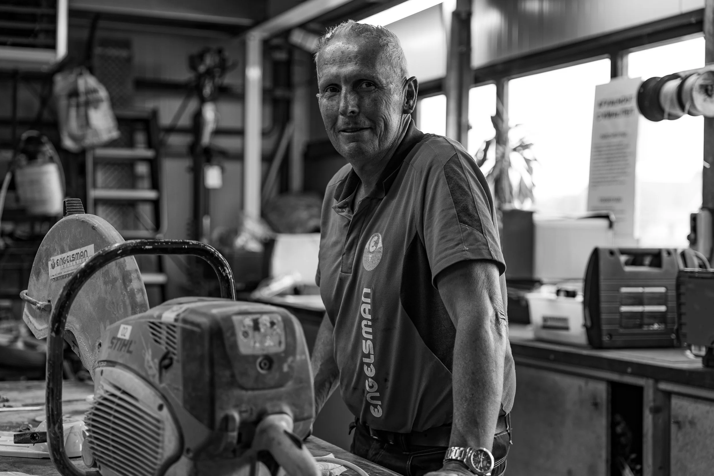 A man in a work shirt standing in a workshop with tools and equipment around him, looking at the camera.