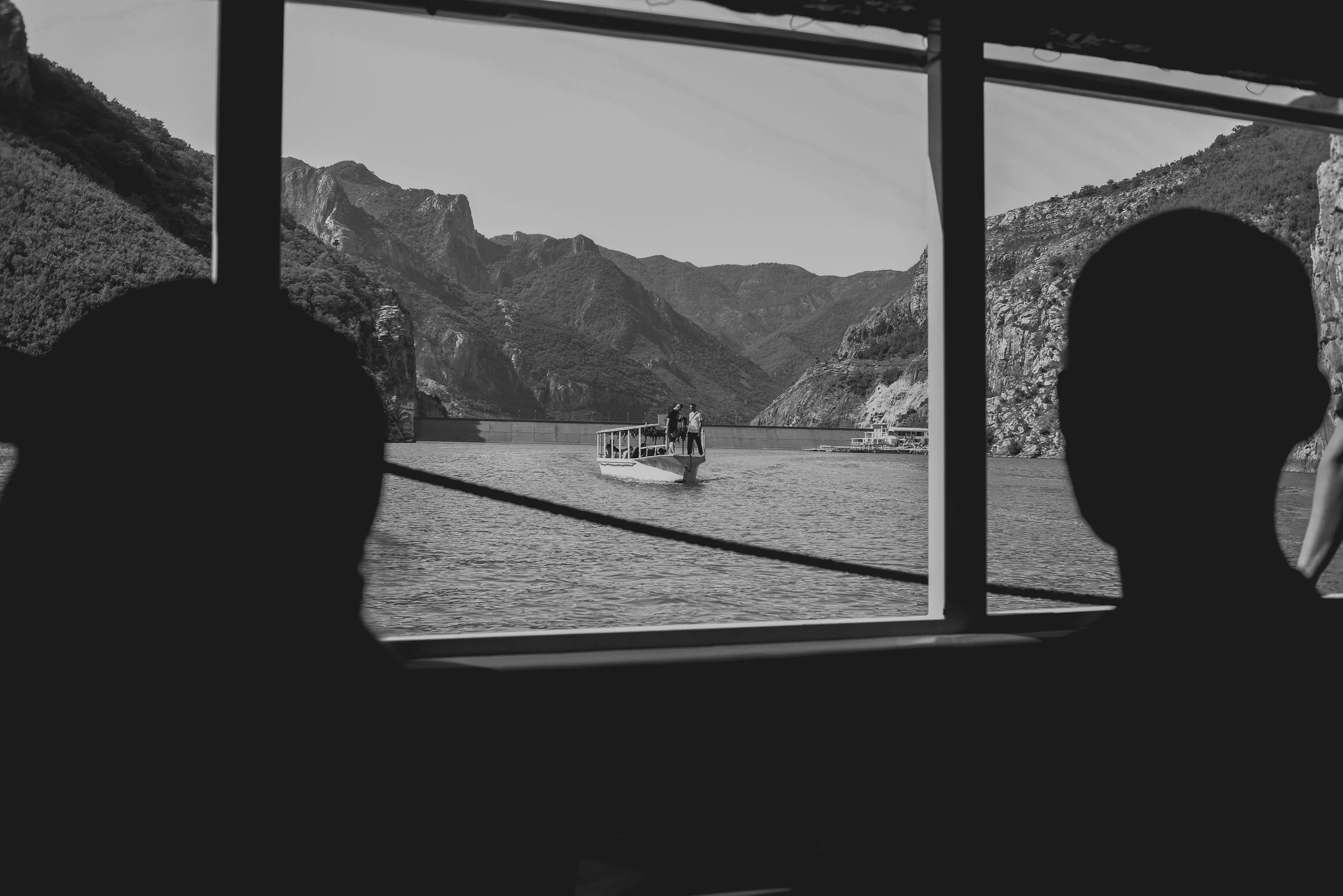 Two silhouetted individuals looking out a window at a large dam with mountains in the background, and a small boat with people on the water.