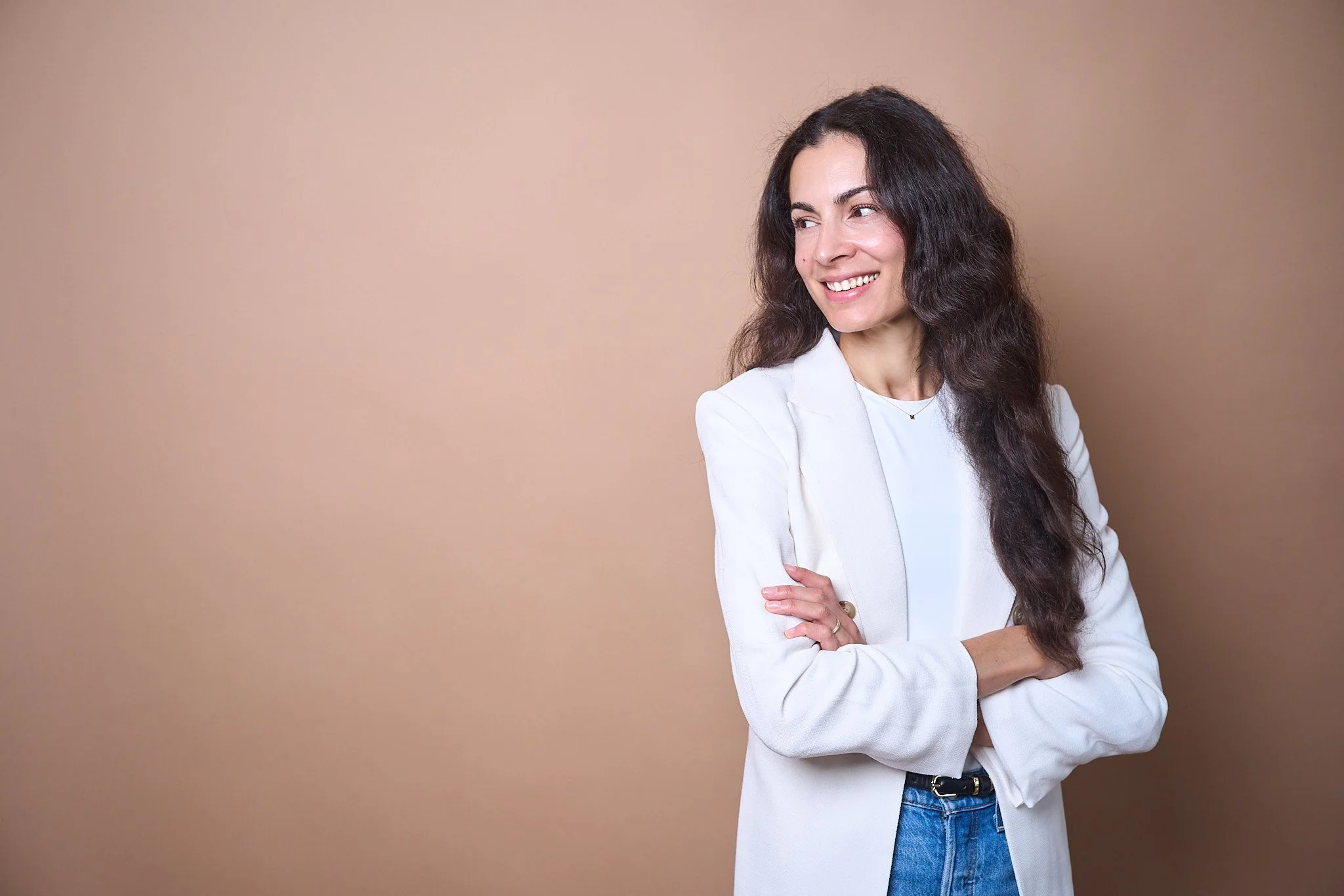 A woman with long dark hair, wearing a white blazer and jeans, smiling and looking to her right, standing against a plain beige background.