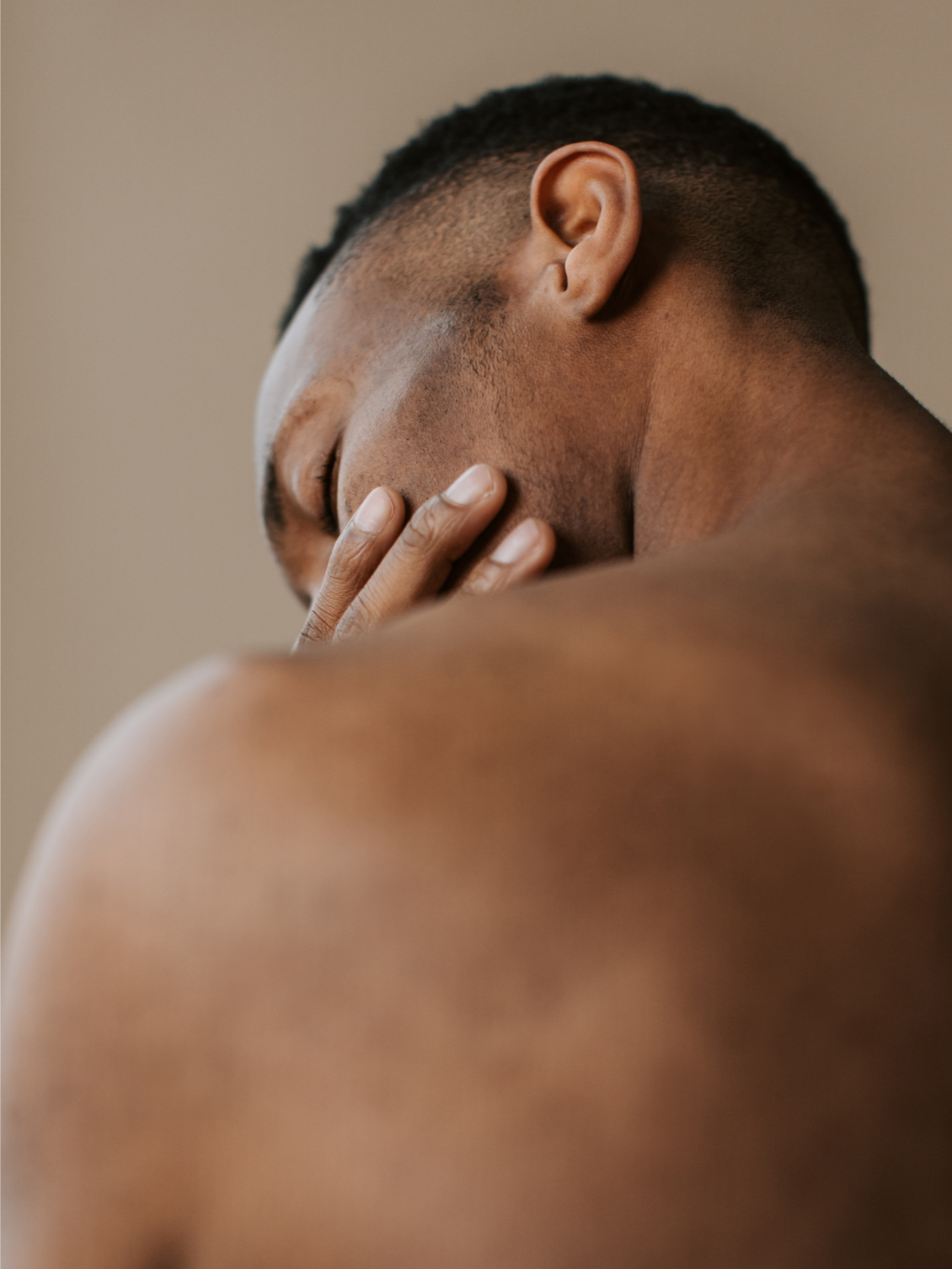 A shirtless man with short curly hair covering his face with his hand, leaning forward.