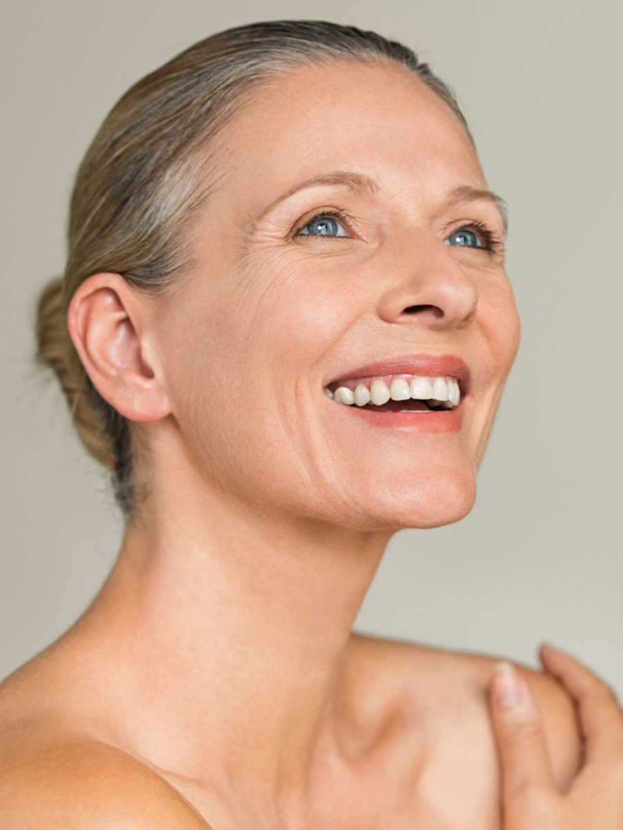 Close-up of a smiling middle-aged woman with light skin, blue eyes, and blonde hair tied back, looking slightly upwards against a neutral background.