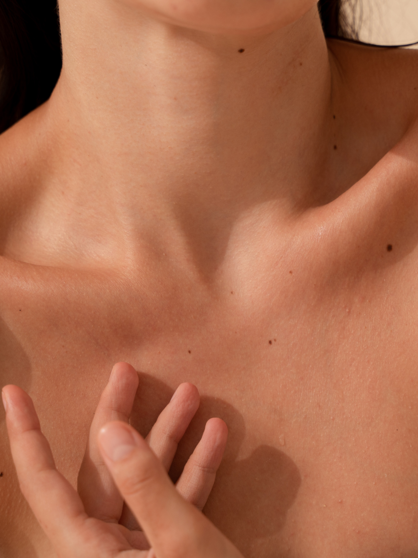 Close-up of a woman's neck, collarbone, and shoulder, with her hand gently touching her collarbone. The skin is smooth and bare, with a few small moles visible.