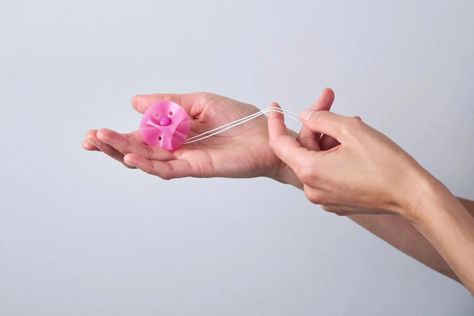 Eine Person hält ein rosa Würfel-Pessar mit weißem Rückholbändchen vor einem schlichten, hellgrauen Hintergrund.
