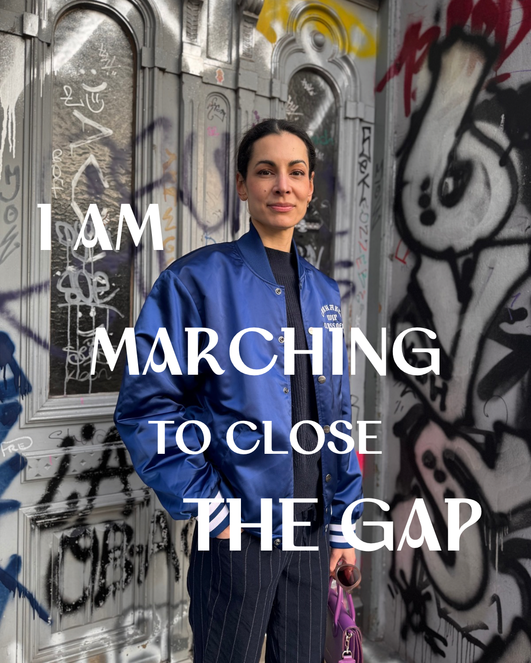 A woman in a blue jacket standing in front of graffiti-covered wall with the text 'I AM MARCHING TO CLOSE THE GAP' overlaid on the image.