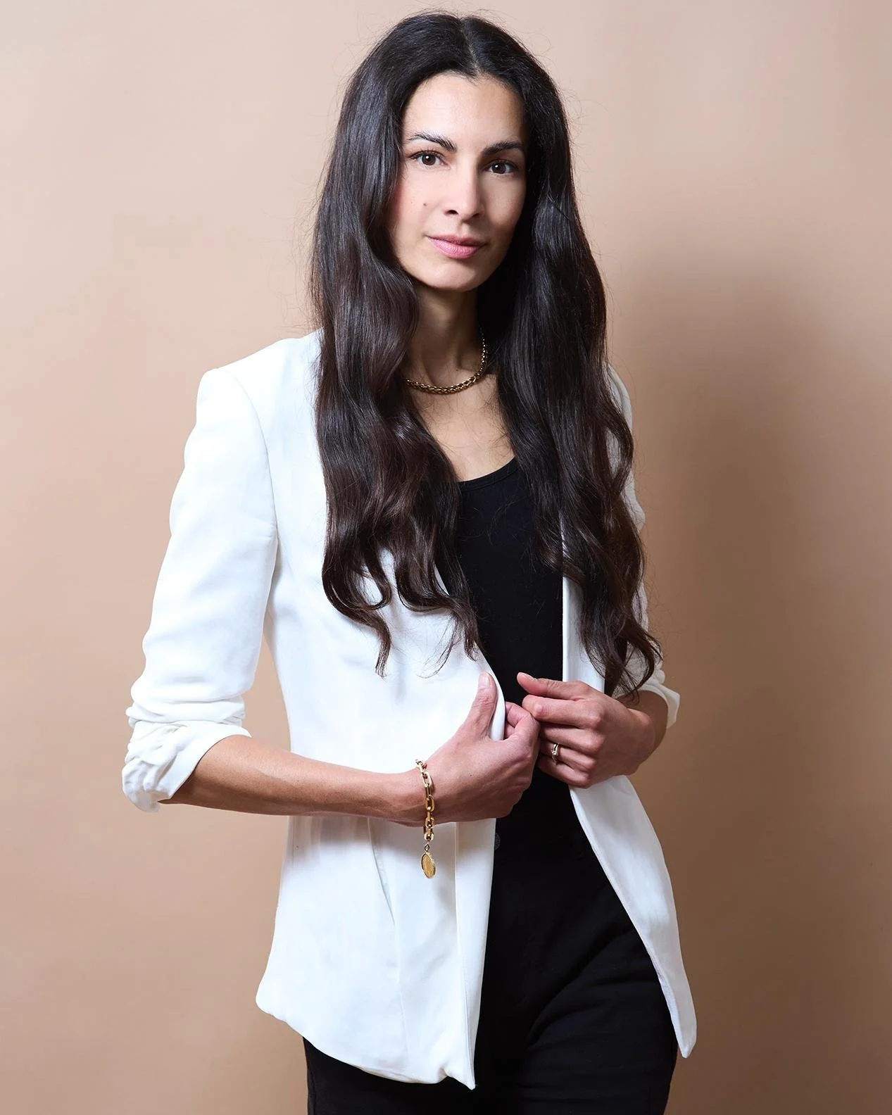 Woman with long dark hair wearing a white blazer and black top, standing against a beige background.