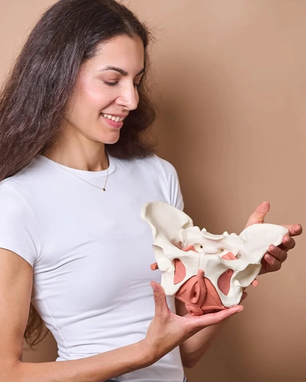 A woman smiling and looking down at a model of a human pelvis that she is holding with both hands.