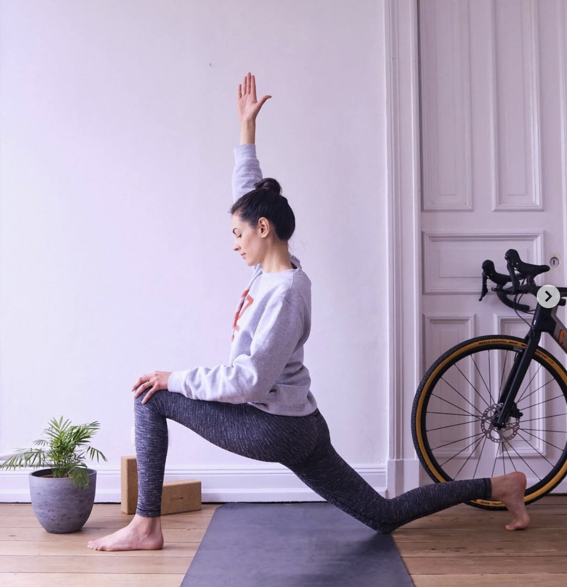 Woman practicing yoga in a lunge pose indoors, with one arm raised and the other on her knee, next to a potted plant, a yoga block, and a bicycle.