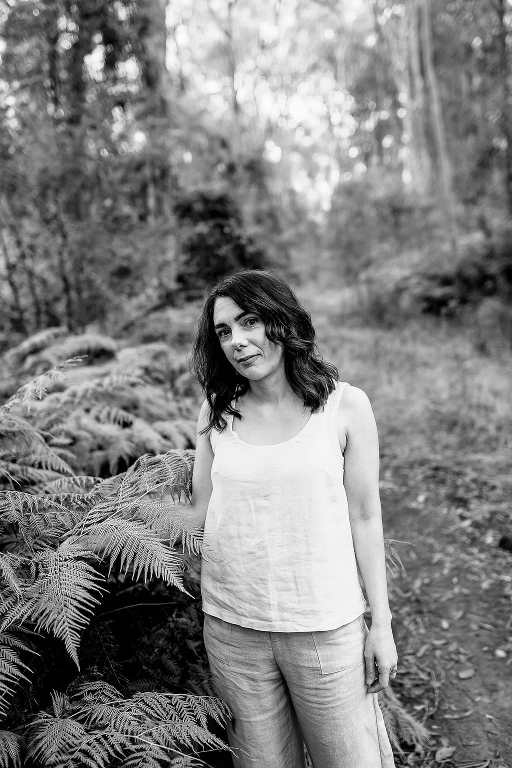 Kylie Riddell Naturopath with dark wavy hair wearing a sleeveless light-colored top and pants standing outdoors among ferns and trees in a blue moutains.