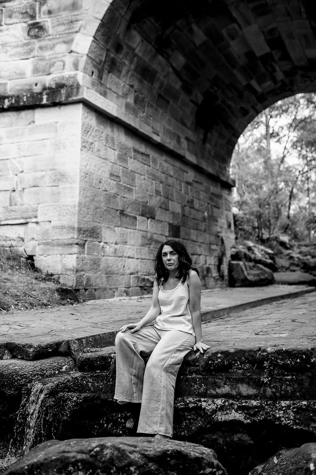 Kylie Riddell Naturopath sitting on a stone ledge under a large stone bridge with an arch, near some rocks and water, in a natural outdoor setting.