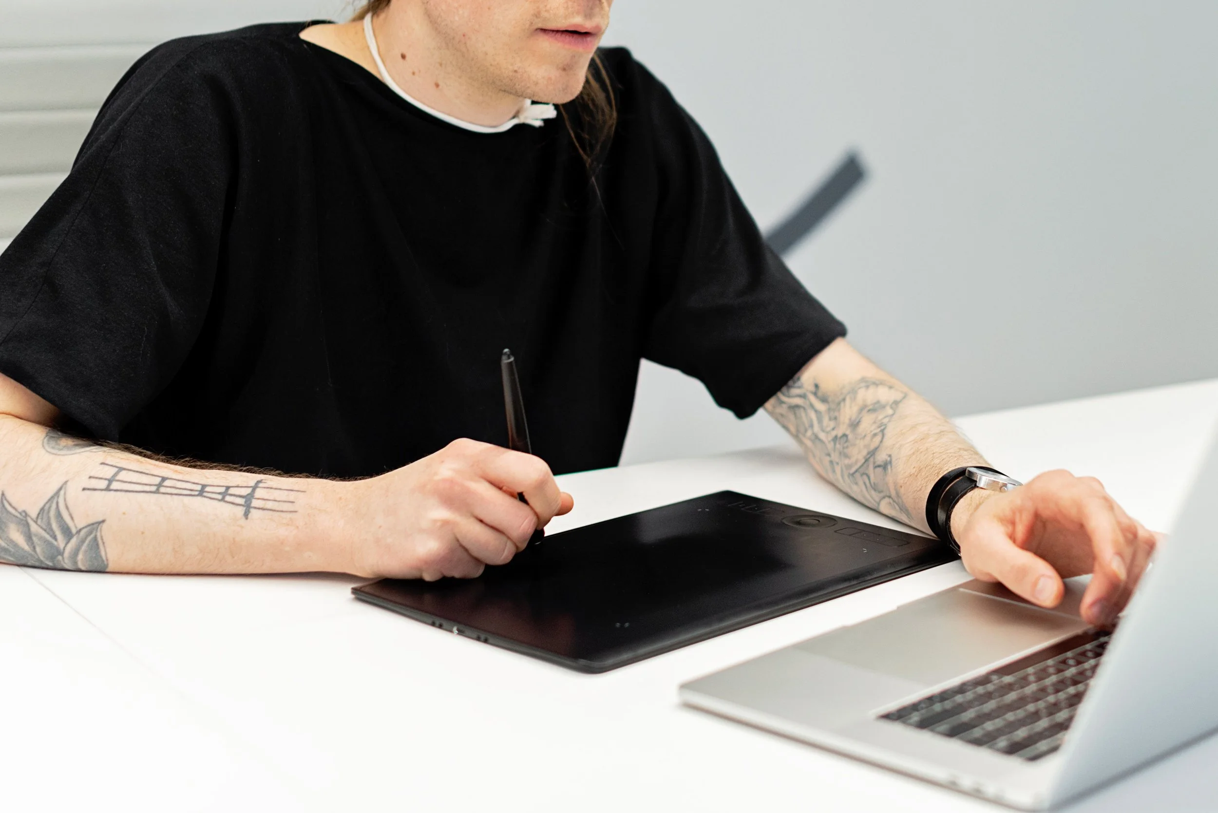A person with tattoos drawing on a tablet, a laptop in front of them.
