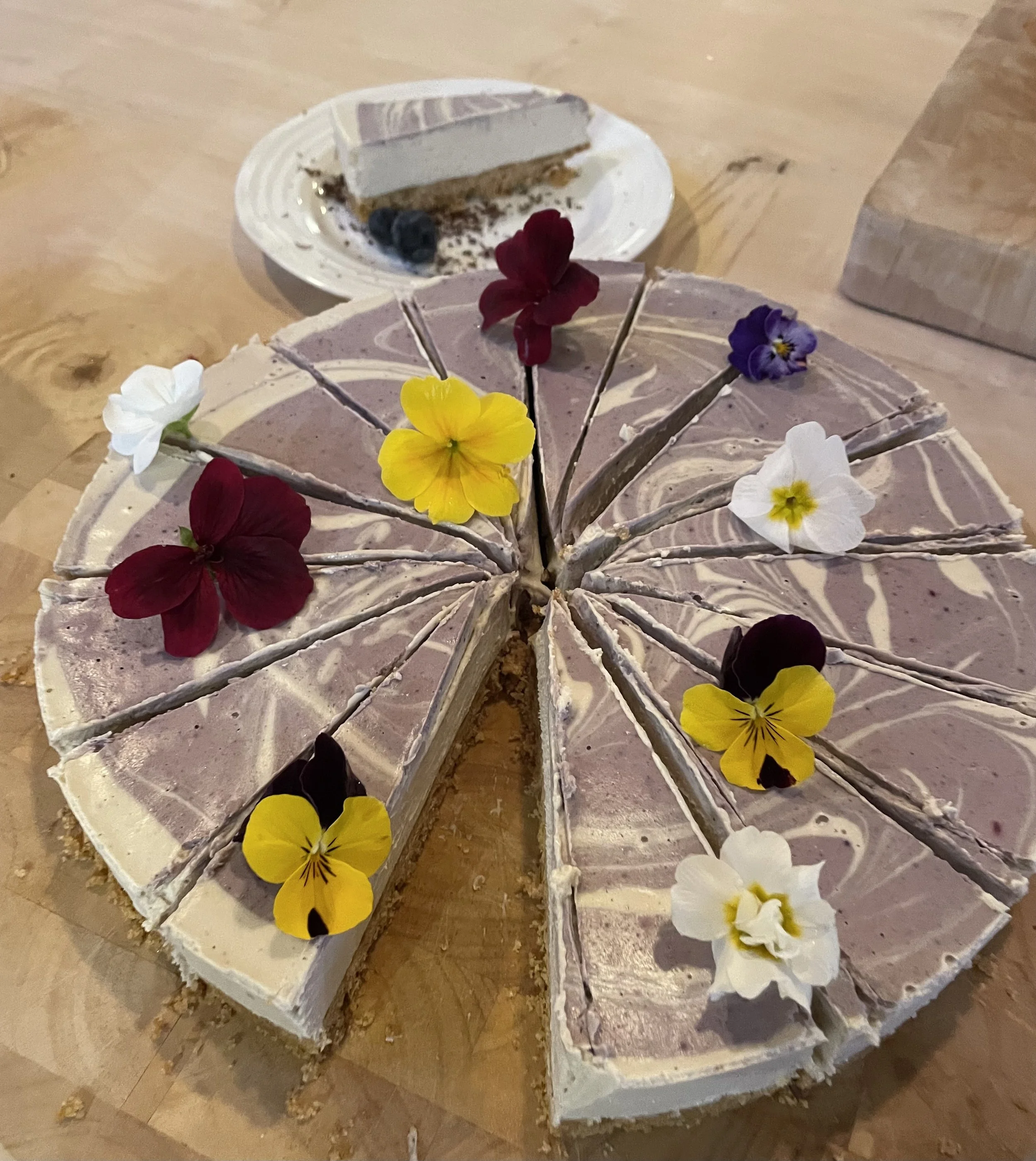 A sliced marble or marble-patterned cake decorated with edible or real flowers, topped with sunflower and pansy flowers, placed on a wooden surface with a piece of cheesecake in the background.