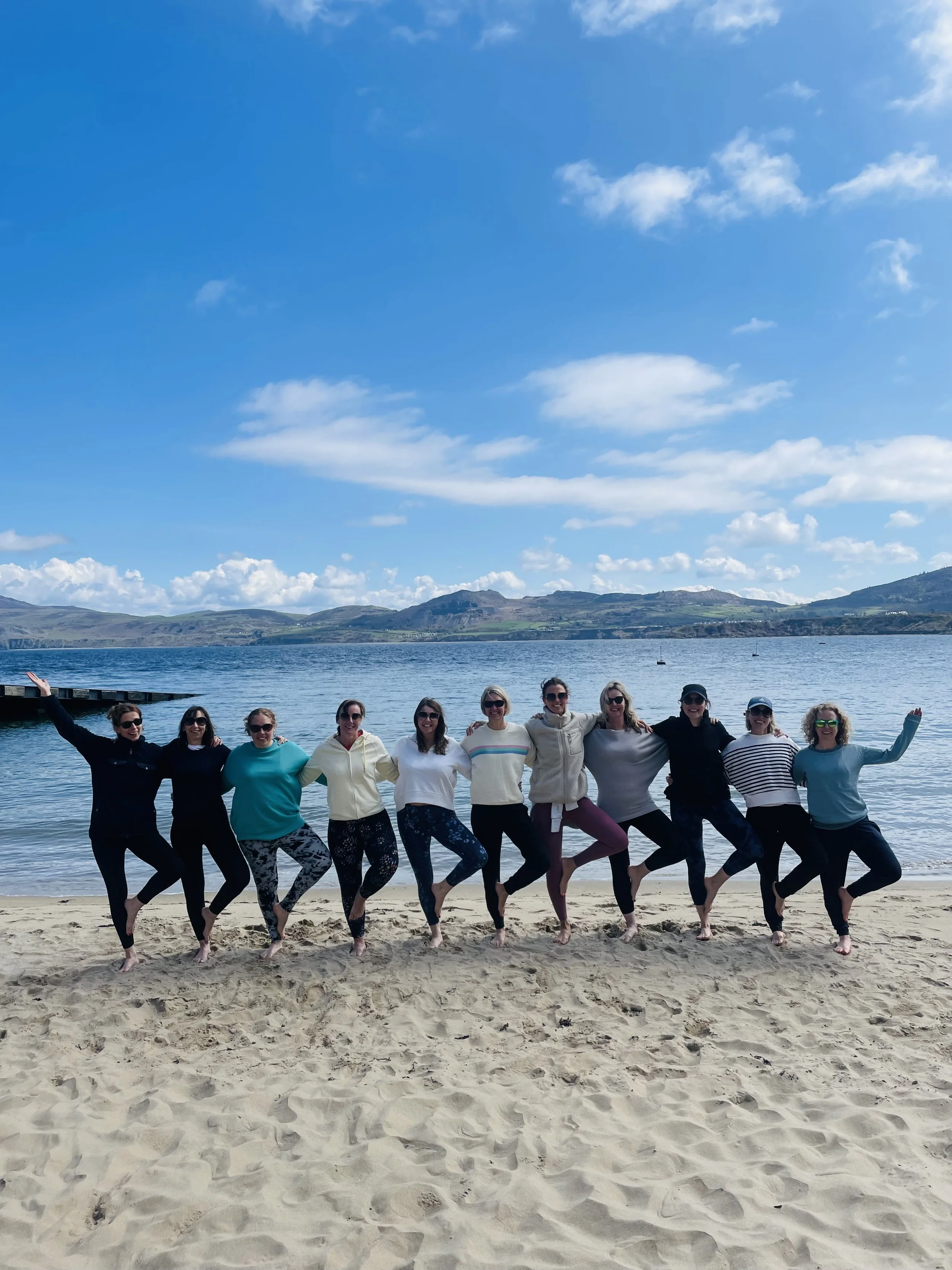 Group of ten women standing on a sandy beach, posing with arms around each other, with water and hills in the background under a partly cloudy sky.
